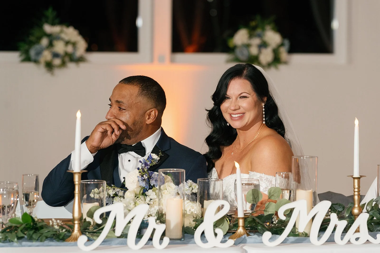 A smiling bride and groom sit at a wedding reception table decorated with candles, flowers, and a 'Mr. & Mrs.' sign. The groom is wearing a tuxedo and the bride is in a wedding dress with a veil. The groom appears to be emotional, covering his mouth 