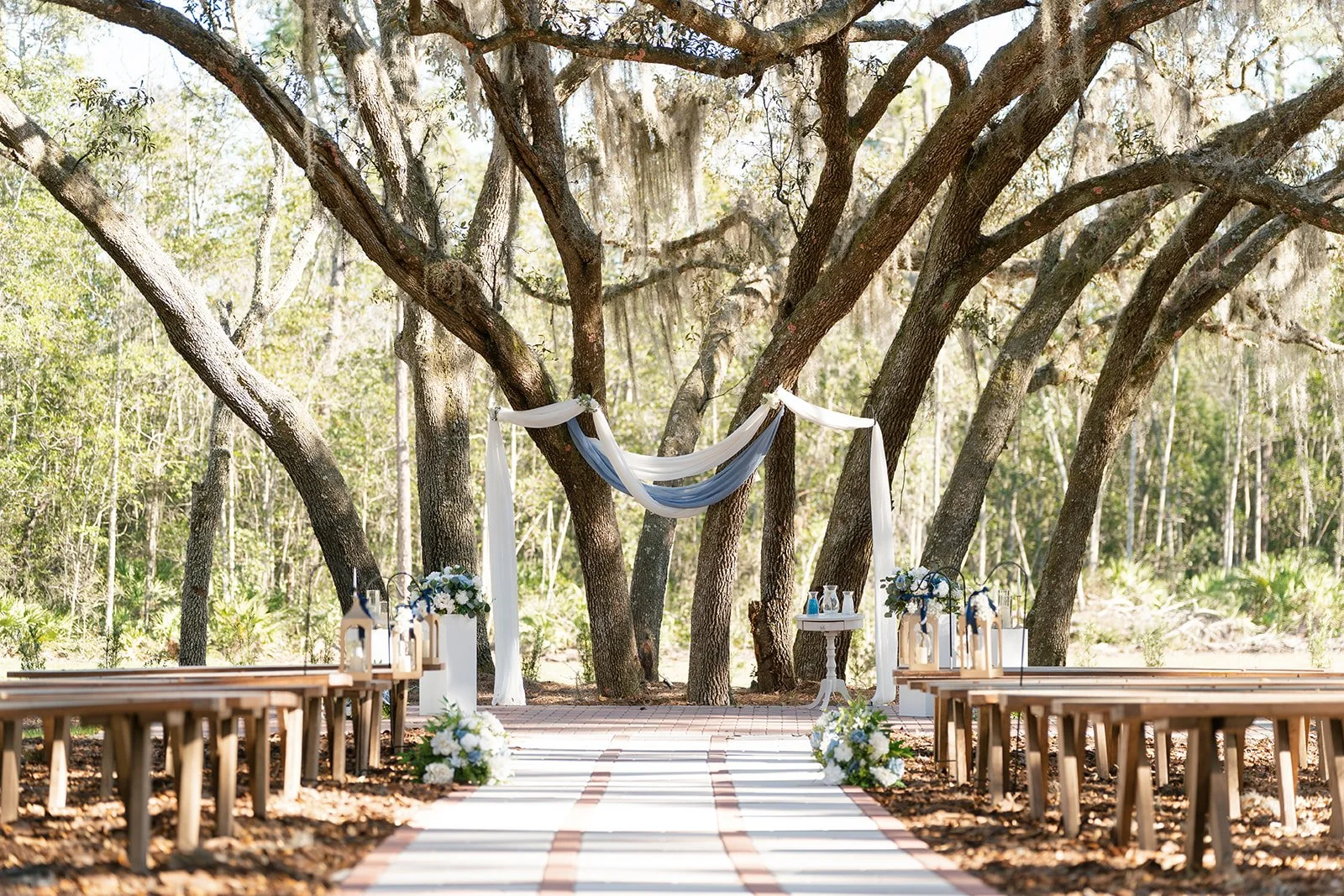 Outdoor wedding setup in a forest with a wooden arch decorated with white and blue fabric, flanked by flower arrangements, and wooden benches on either side.