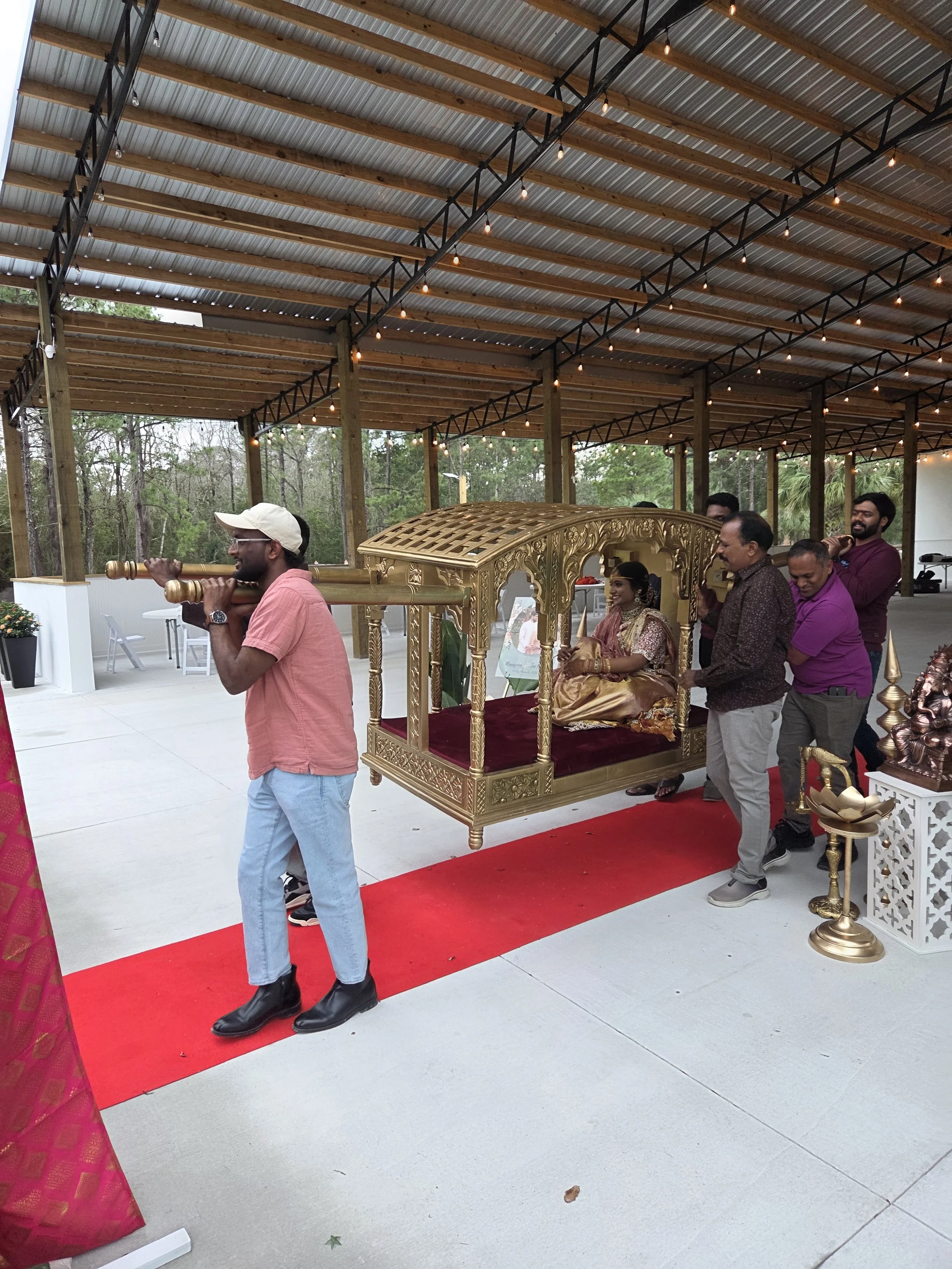 People participating in an Indian wedding ceremony, with a bride seated on a decorated palanquin, being carried by men, under a covered outdoor area with string lights.