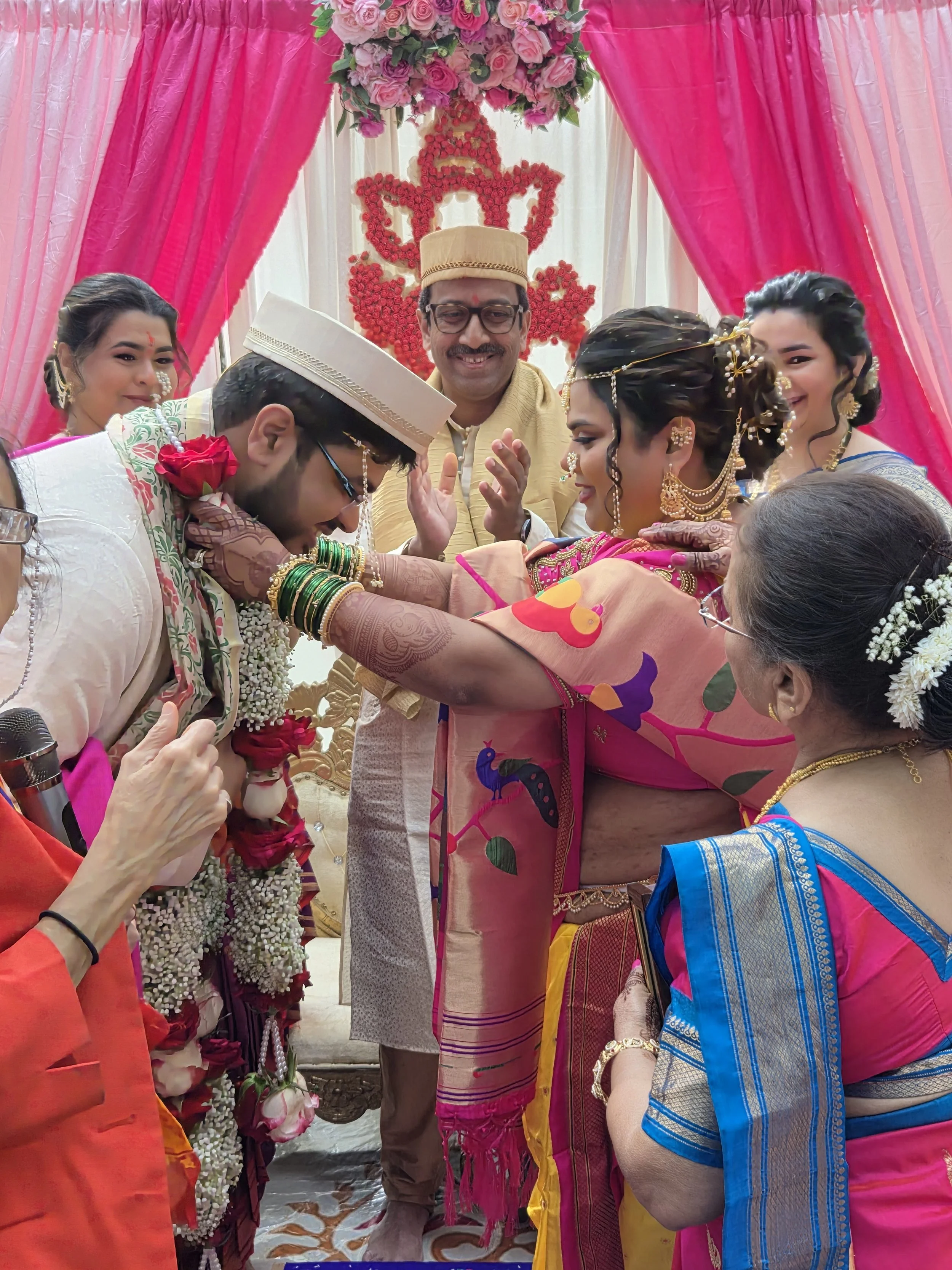 A group of people at a traditional Indian wedding ceremony. The bride dressed in a pink saree with gold jewelry, and the groom dressed in a cream-colored sherwani with green bangles. The family stands under a decorated pink and cream wedding canopy w