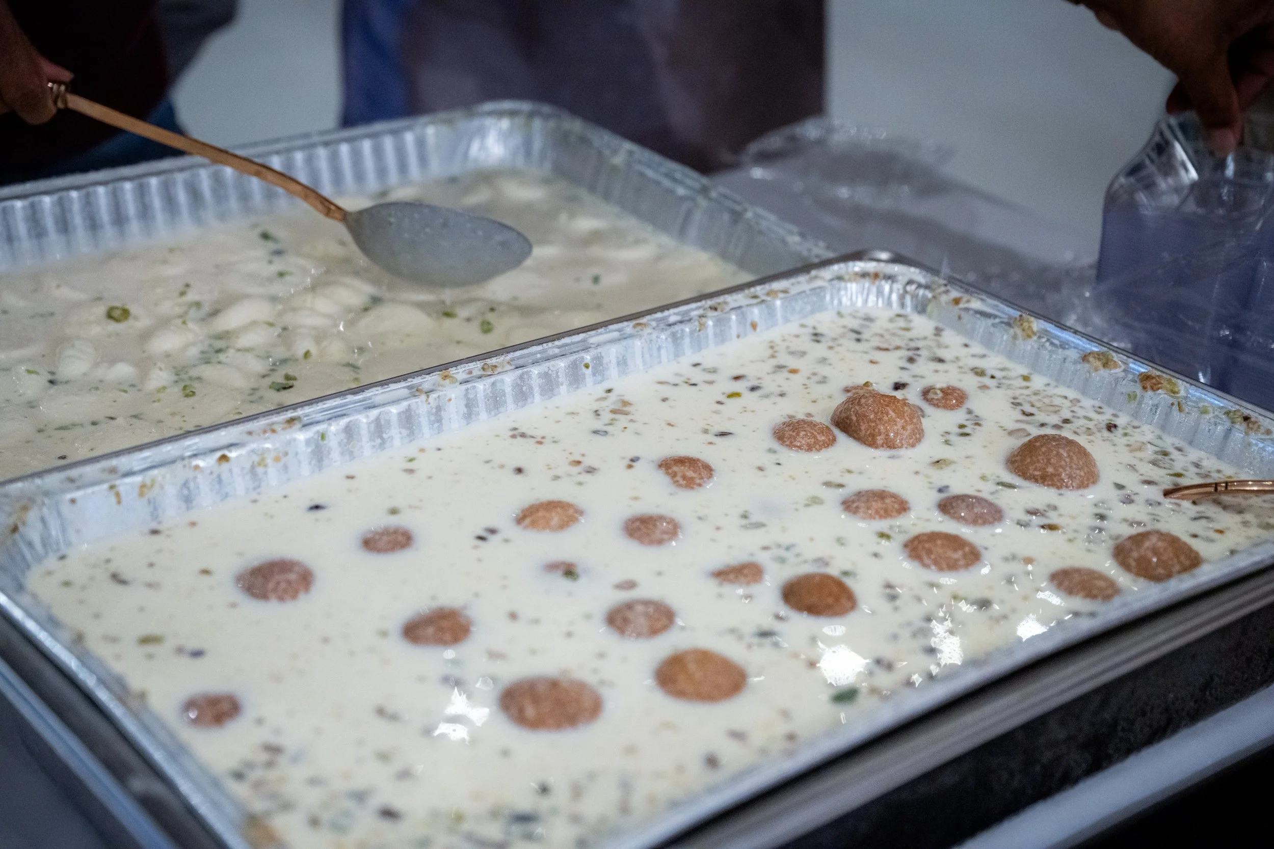 Two aluminum trays filled with a creamy dessert, possibly containing small green and dark-colored mix-ins, and topped with round chocolate or cookie pieces. Someone is serving the dessert with a spoon.