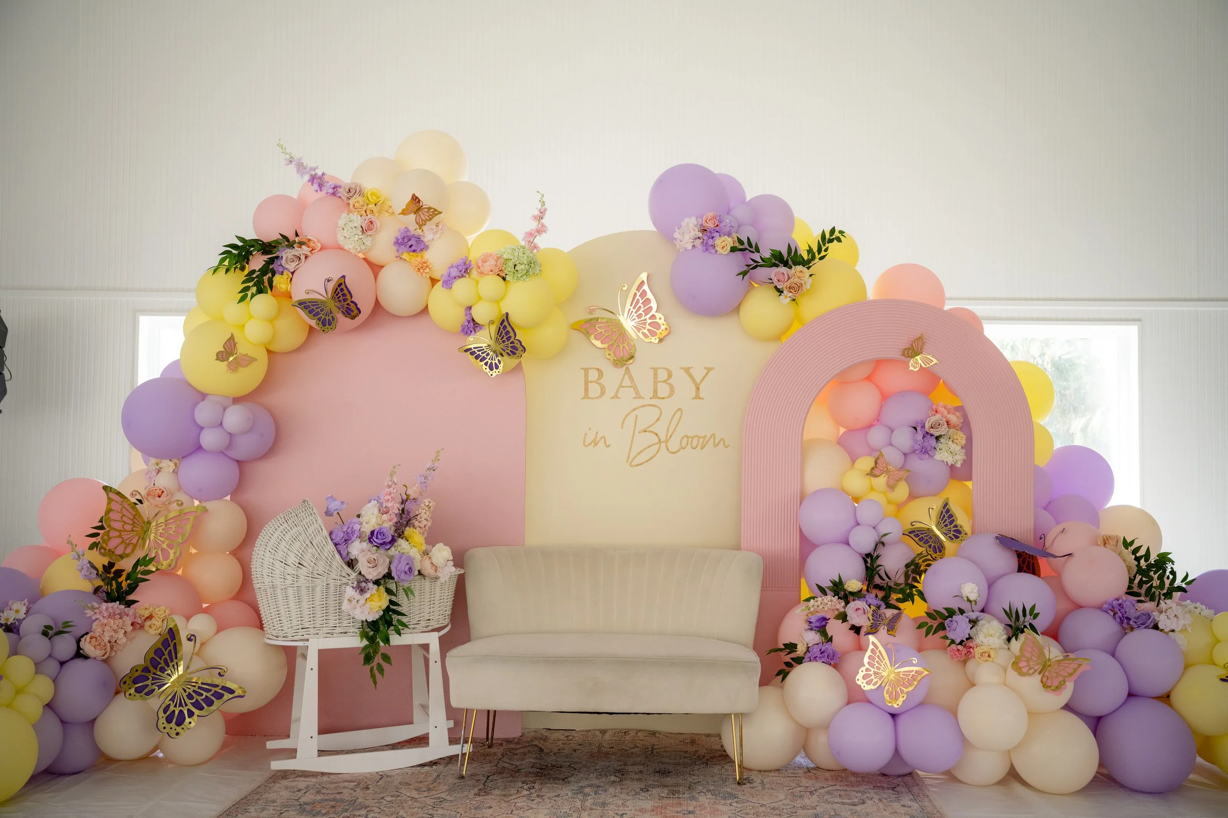 Decorative backdrop for a baby shower with pastel-colored balloons, floral arrangements, and butterfly ornaments. A sign reads 'Baby in Bloom.' There is a white chair and a white cushioned bench in front of the display.