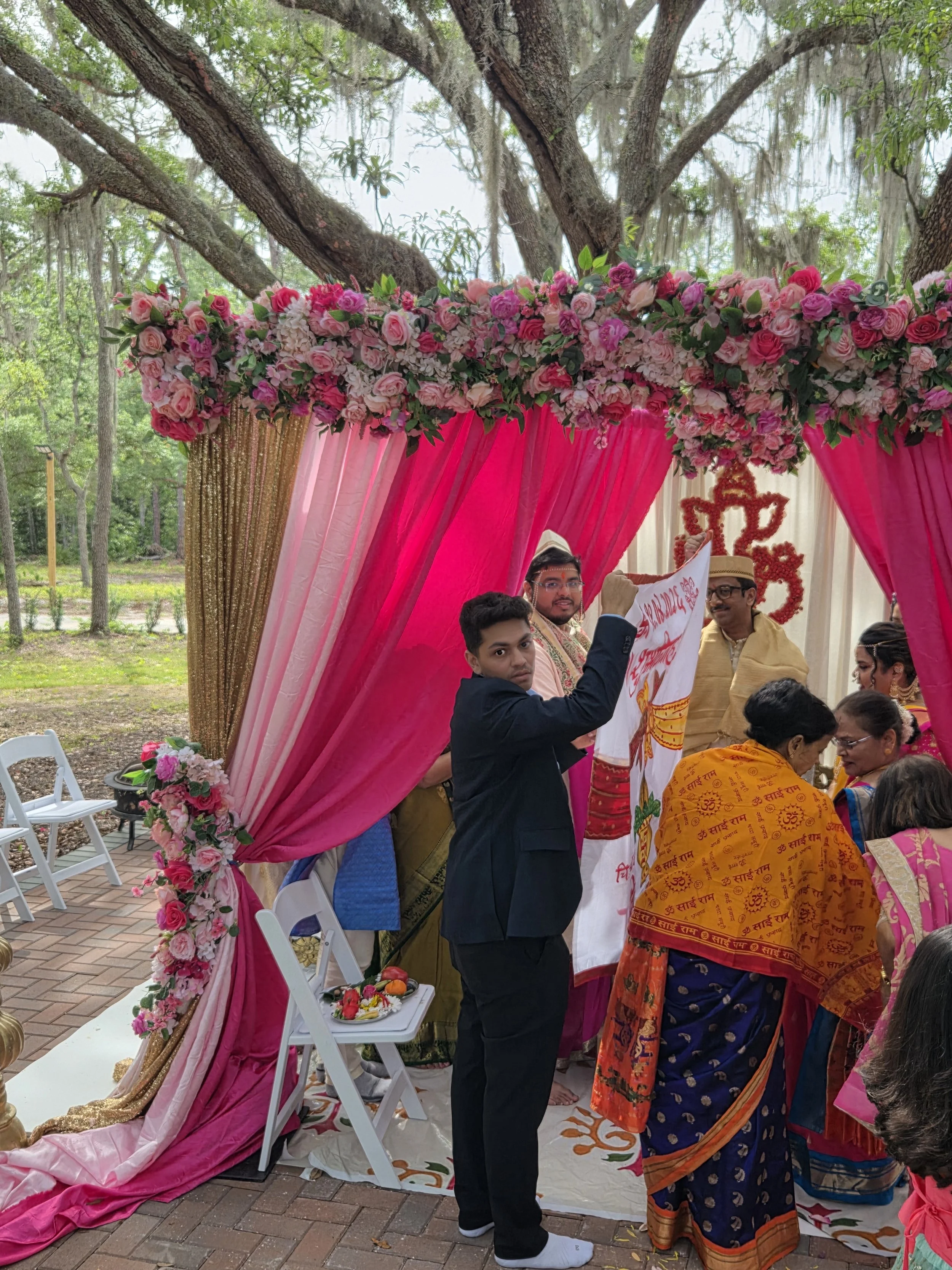 This image depicts a traditional Indian wedding ceremony. Several people are gathered under a decorated canopy with pink and gold fabric, surrounded by flowers. The group includes men and women dressed in colorful traditional Indian attire. One man i