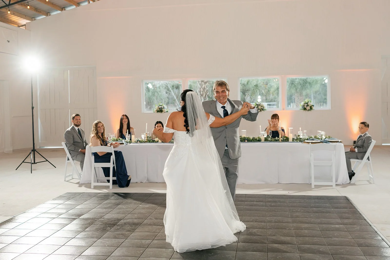 A bride and groom sharing their first dance during a wedding reception, with six seated guests at a long head table behind them.