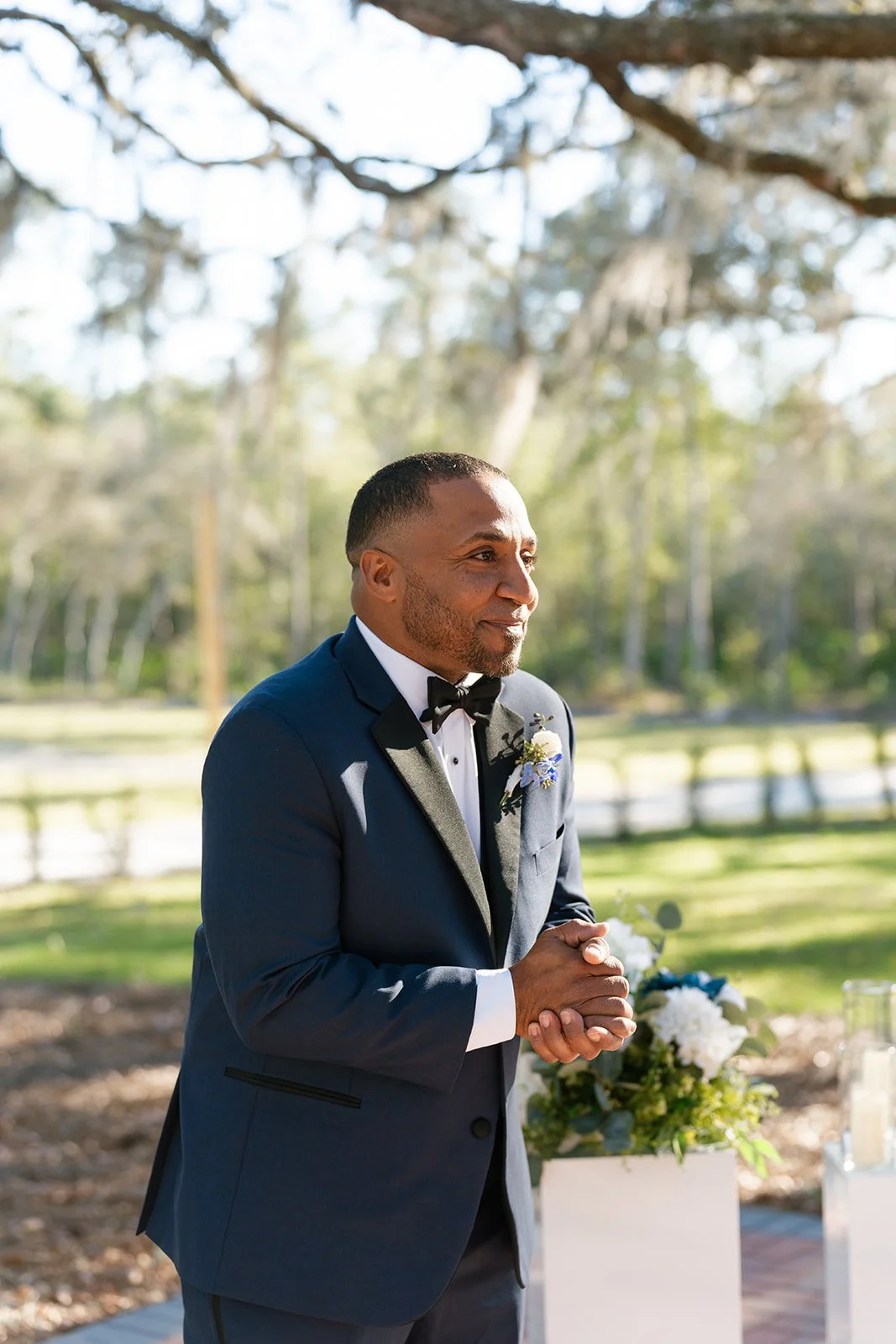A man in a dark suit with a bow tie, standing outdoors during a sunny day, with his hands clasped and smiling softly, next to a floral arrangement.