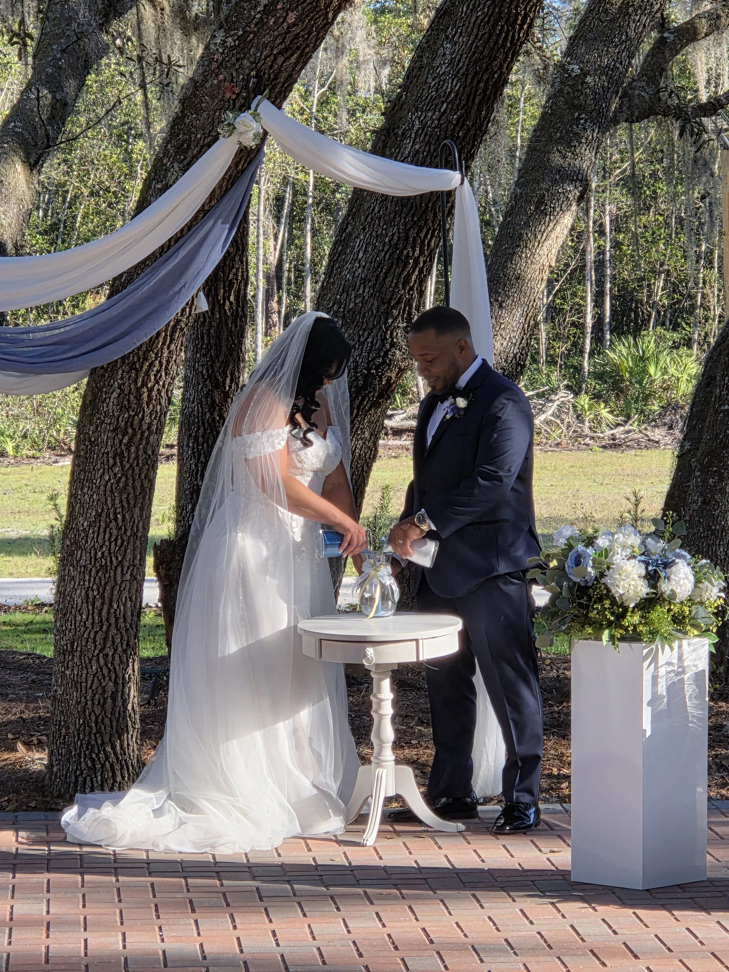 A bride and groom exchange vows during their outdoor wedding ceremony under trees, with white and navy fabric draped from the trees and floral arrangements nearby.