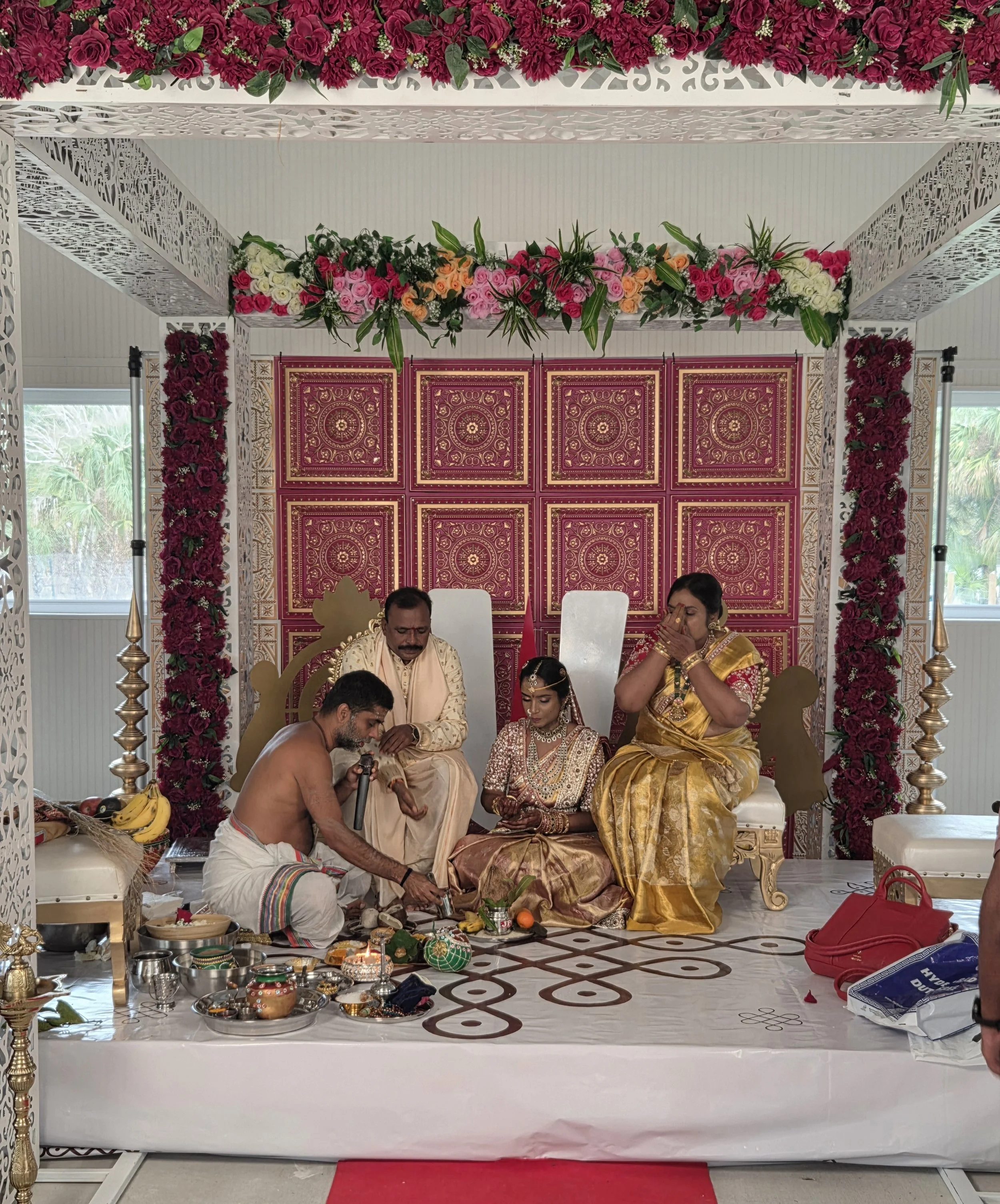 Indian wedding ceremony with a bride and groom performing a ritual on a decorated stage with floral arrangements and traditional items.