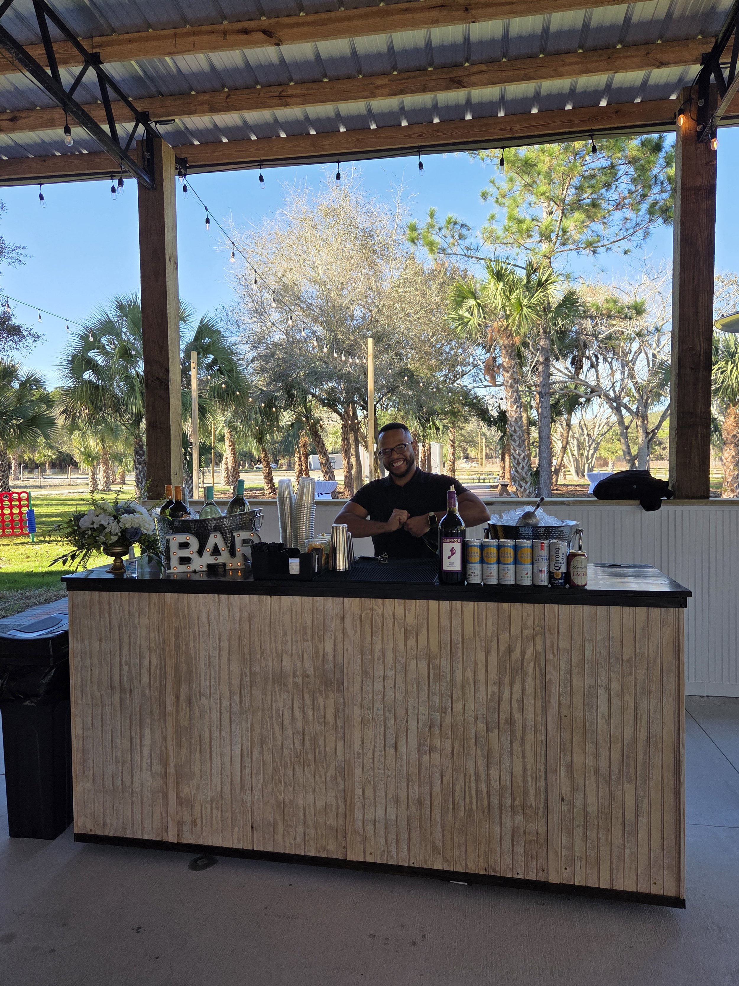 A man standing behind a bar at an outdoor event with trees and string lights in the background. The bar has a sign that says 'BAR' and various bottles, cups, and decorations on top.