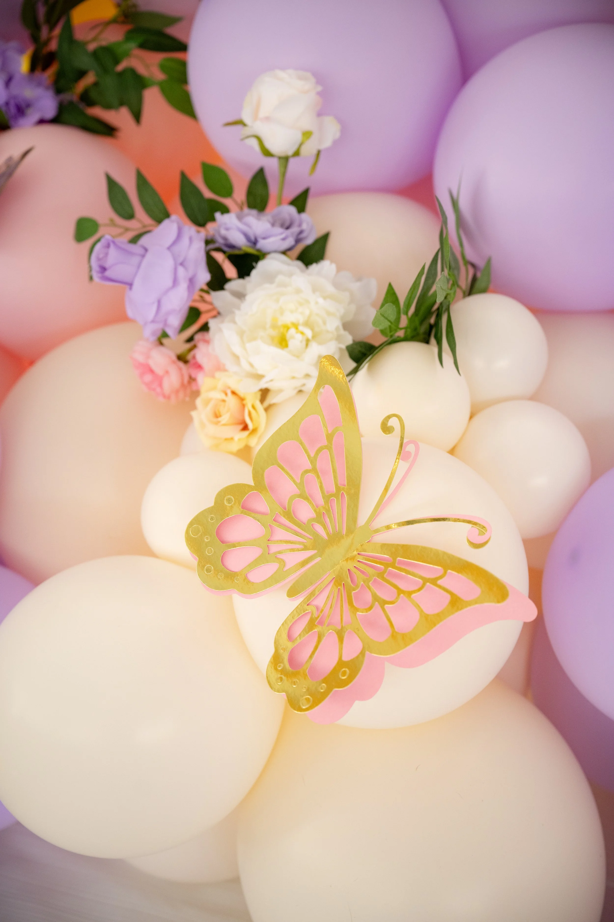 A close-up of pastel-colored balloons, pink, purple, and cream, decorated with artificial flowers and a pink and gold butterfly ornament.