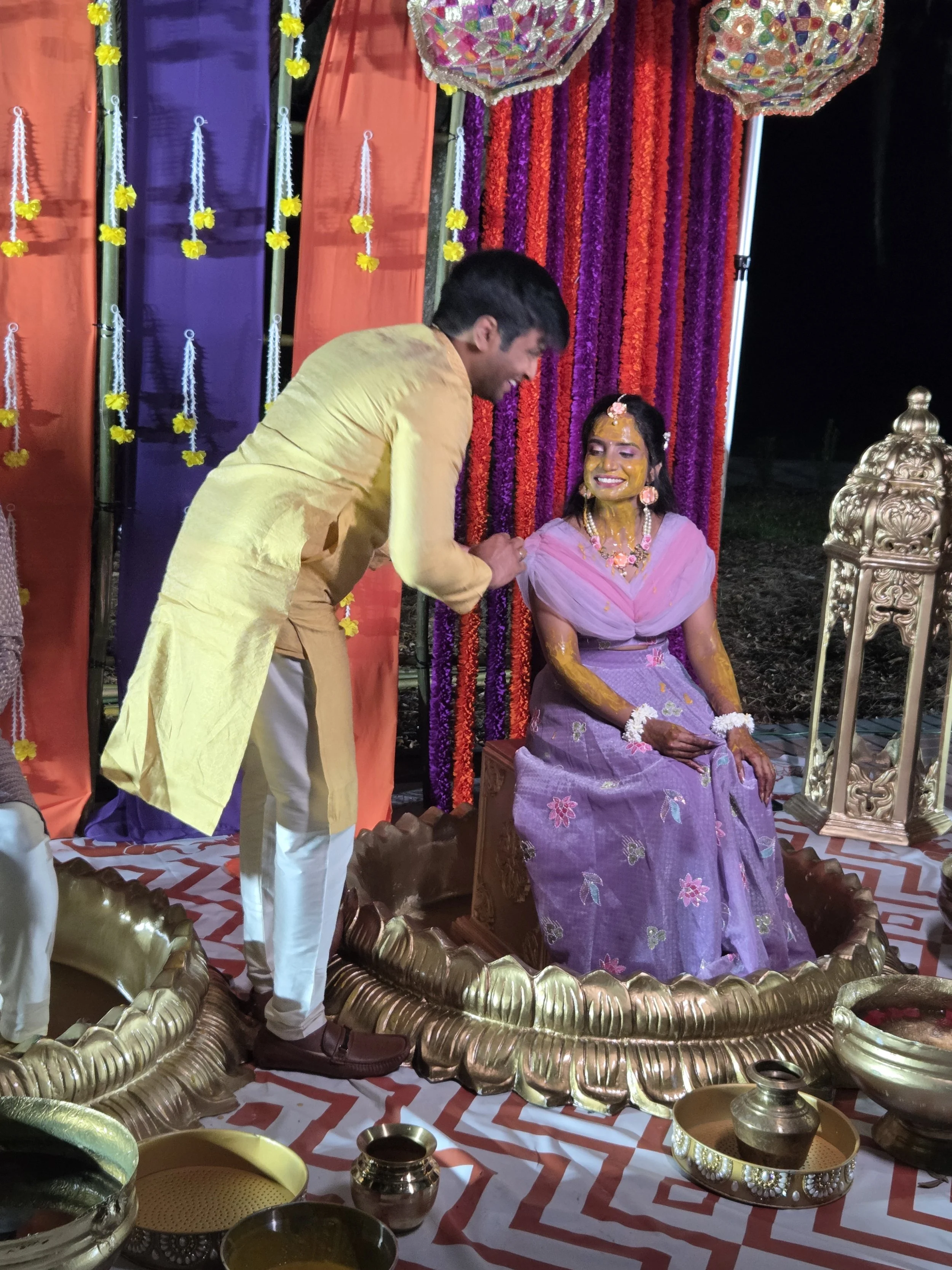 A traditional Indian ceremony where a man is applying a yellow paste to a woman sitting on an ornate golden throne. The woman is dressed in purple, adorned with jewelry, and has flowers on her hair. The background features colorful curtains and flora