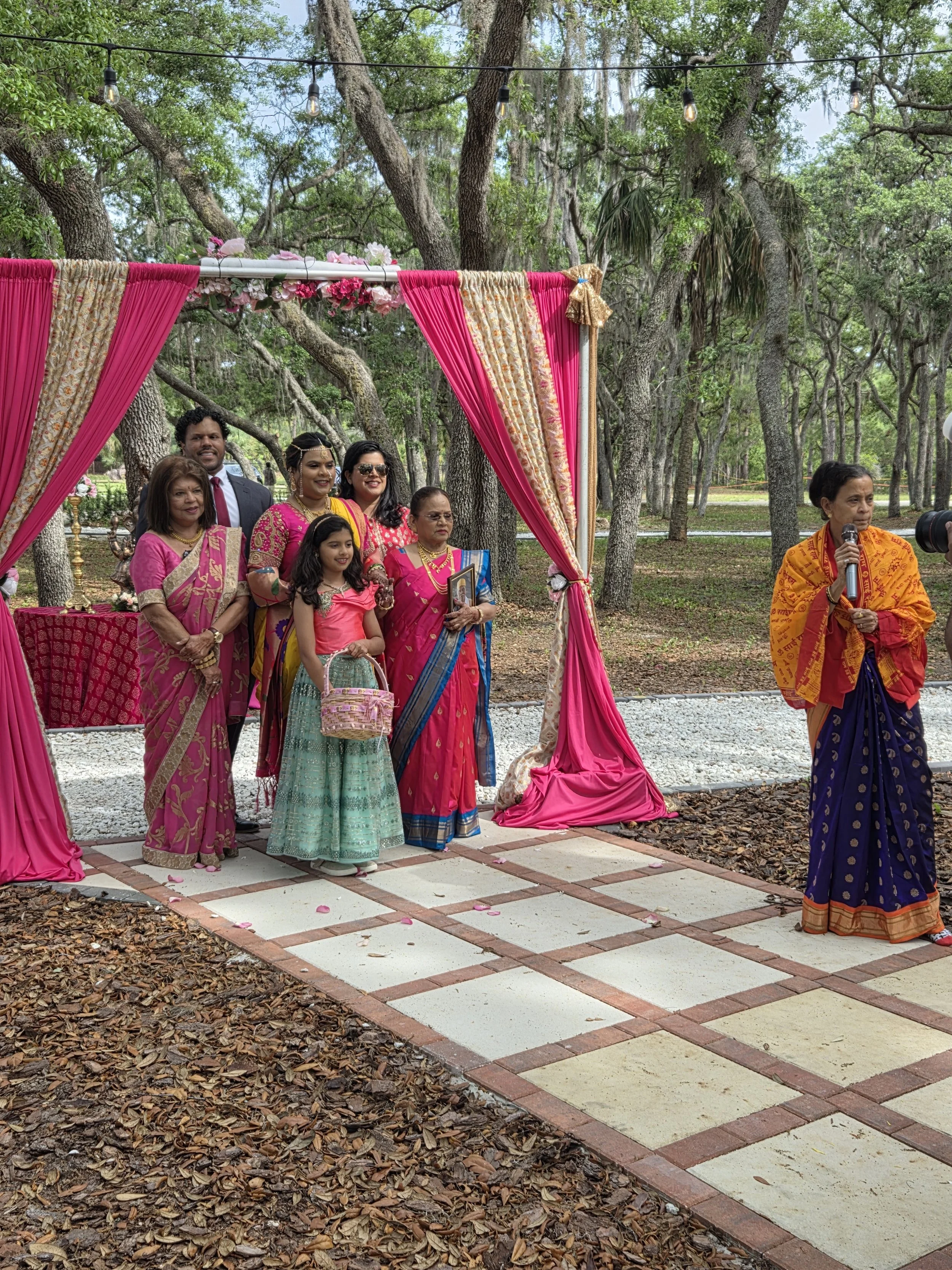 A group of people dressed in traditional Indian clothing at an outdoor wedding ceremony with a pink and gold decorated canopy in a wooded park.