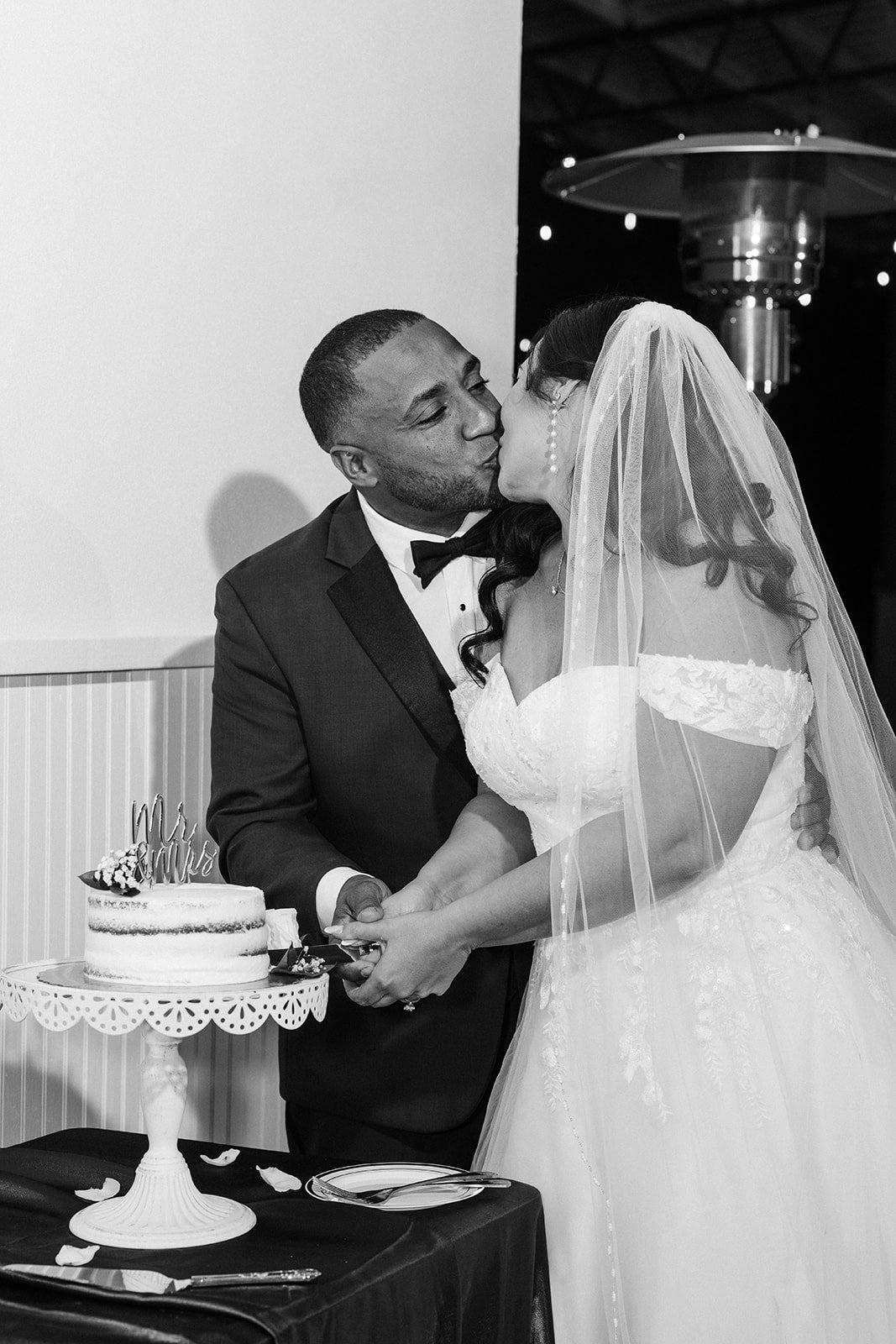 A groom and bride in wedding attire kissing while cutting a wedding cake at reception.