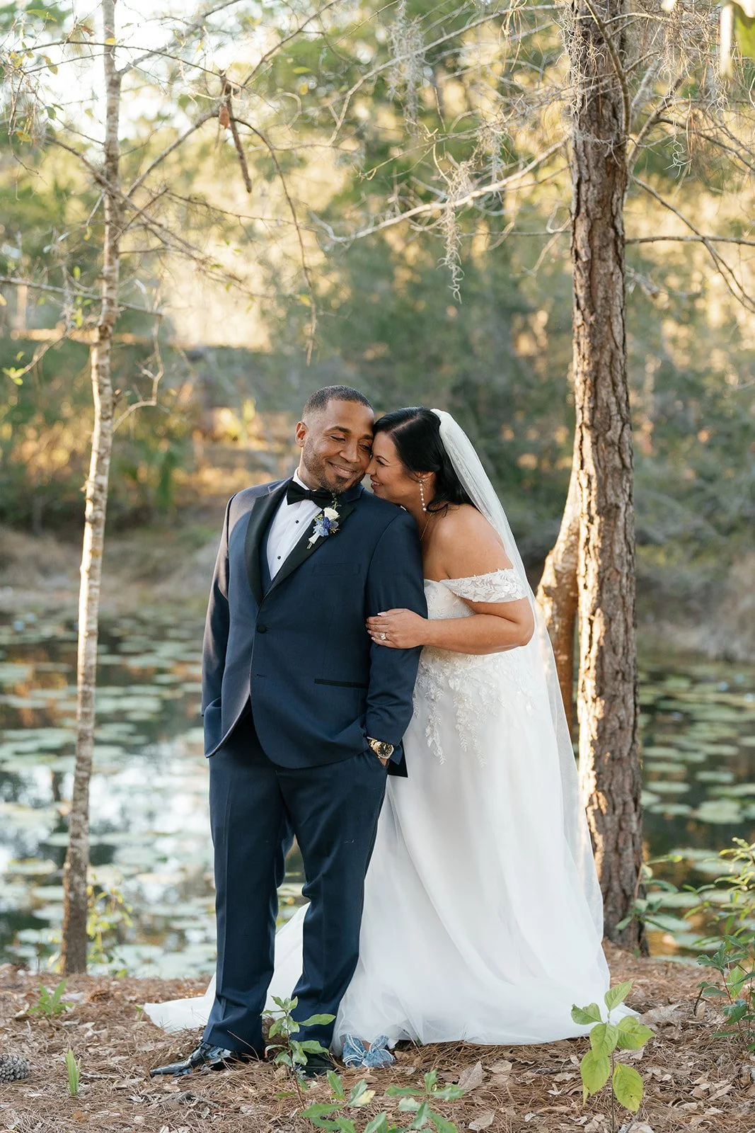 A newlywed couple, a man in a navy tuxedo and a woman in a white wedding dress, sharing a joyful moment outdoors near a pond in a forest with trees and lily pads, during sunset.