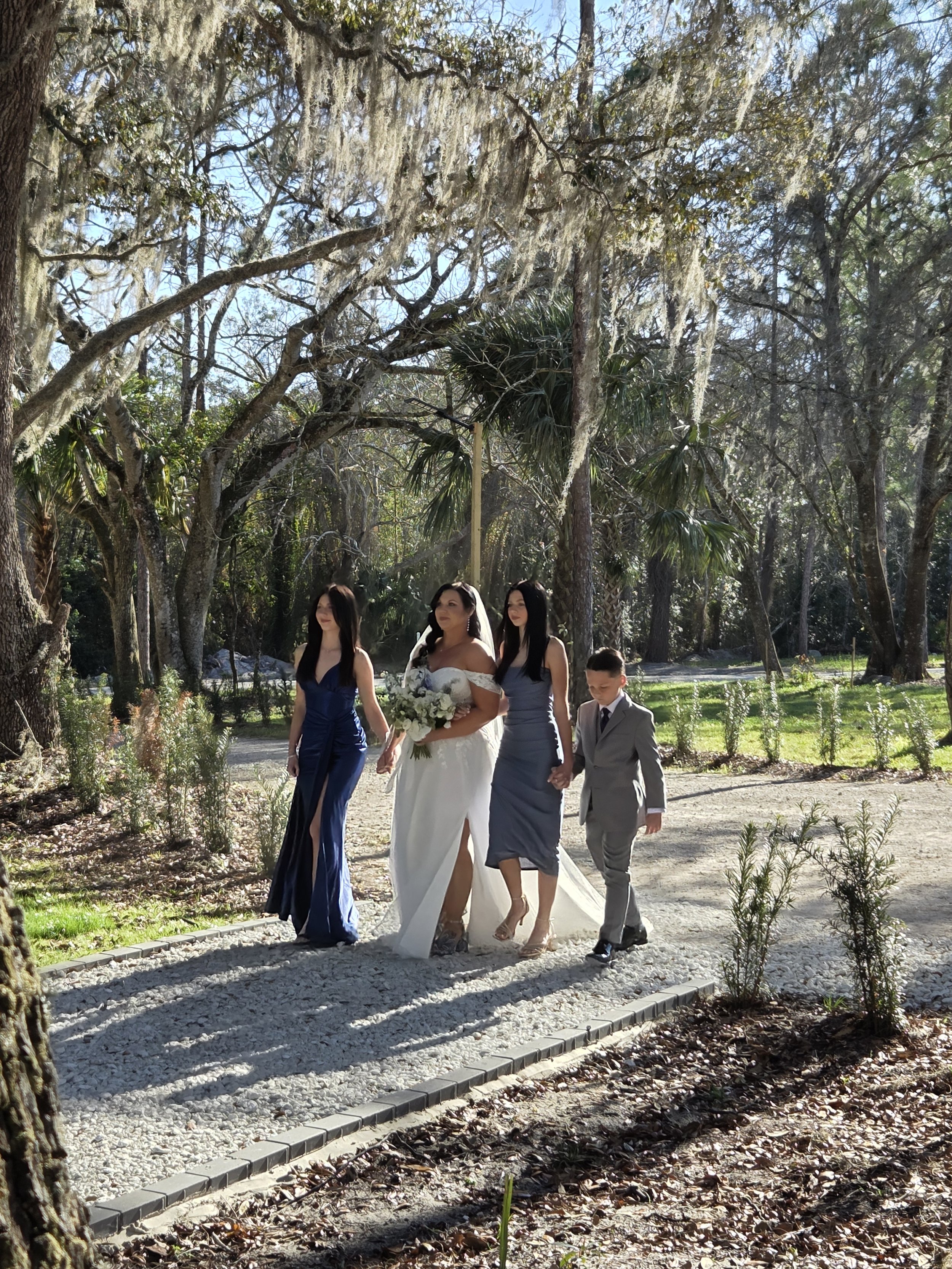 A group of people walking outdoors in a natural setting with trees and greenery, including a woman in a white dress holding a bouquet, accompanied by a woman in a blue dress, a woman in a gray dress, and a young boy in a gray suit.