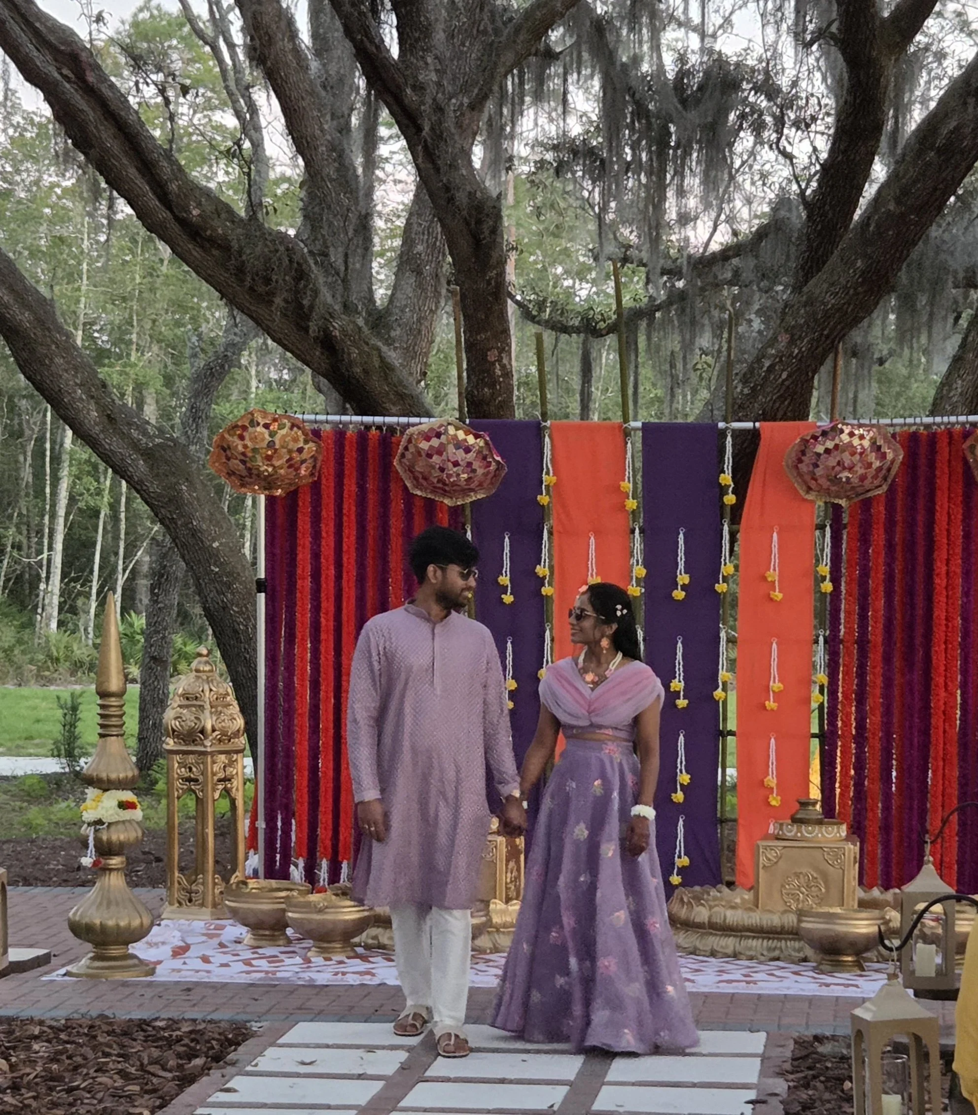 A couple dressed in traditional Indian attire, standing holding hands during a cultural or wedding celebration outdoors. Behind them, a decorated stage with drapes in purple and orange, hanging marigold garlands, and large decorative lanterns. Large 