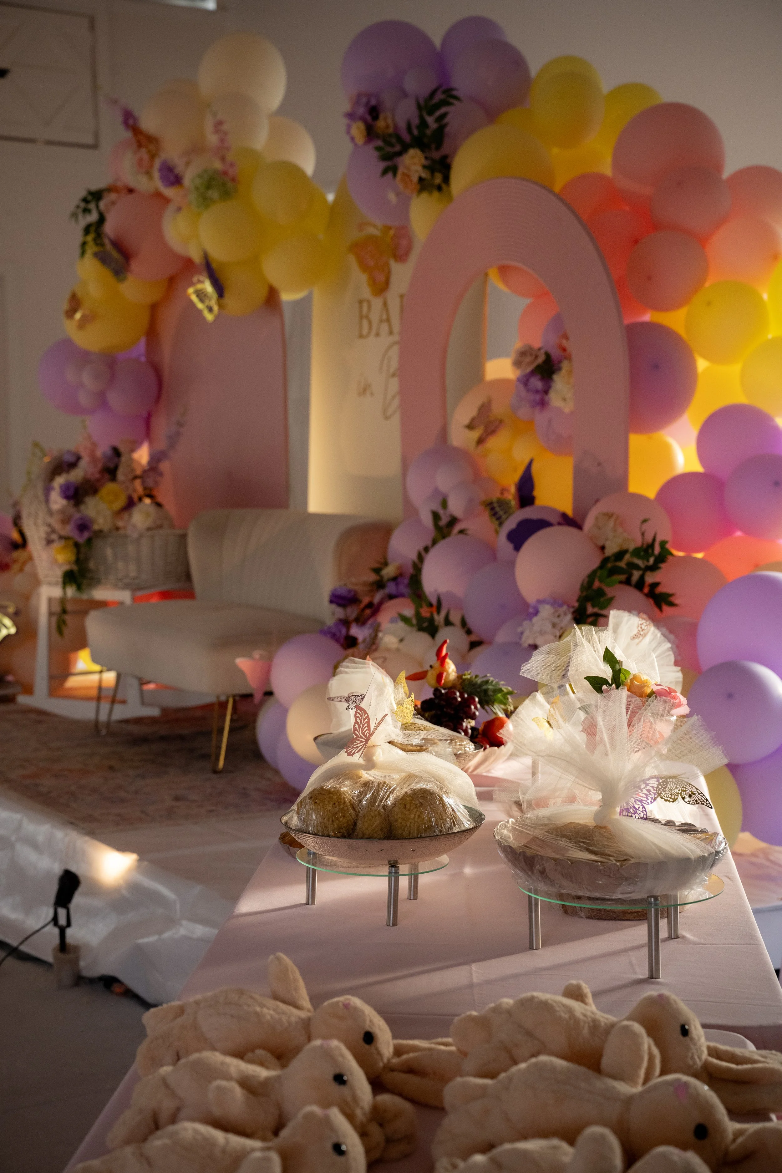 Decorative celebration setup featuring balloon arrangements in pastel colors, a table with stuffed animal toys, and a backdrop with flowers and balloons.