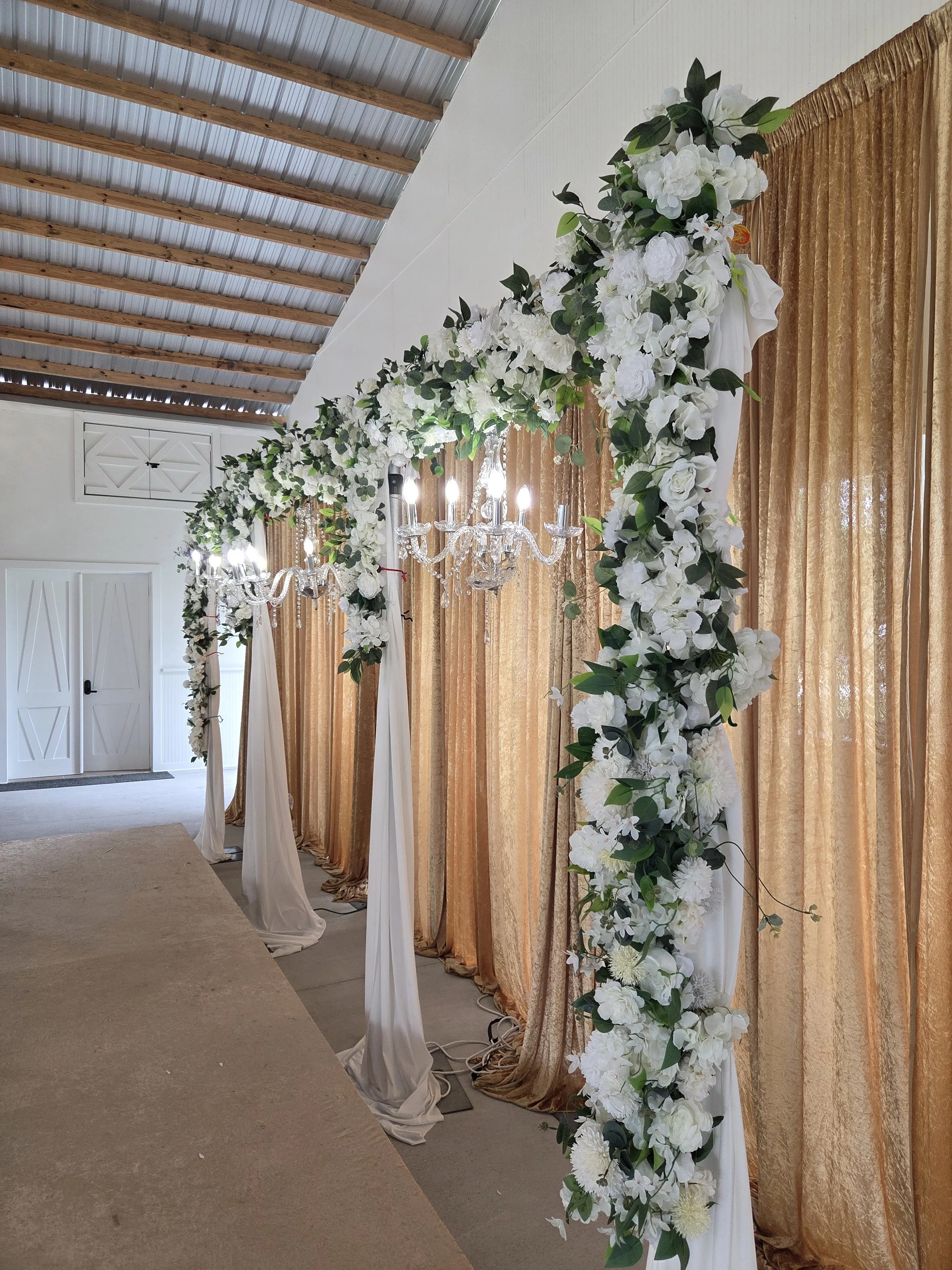 A decorated wedding arch with white flowers and green leaves, white fabric drapes, golden curtains in the background, and crystal chandeliers hanging from the ceiling in an indoor setting.