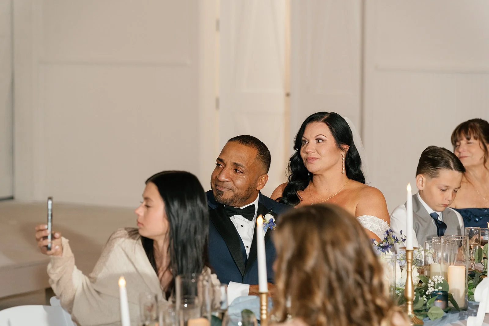 Group of wedding guests seated at a decorated table, including a man in a tuxedo with a bow tie and a woman in a strapless dress, attending a wedding reception.
