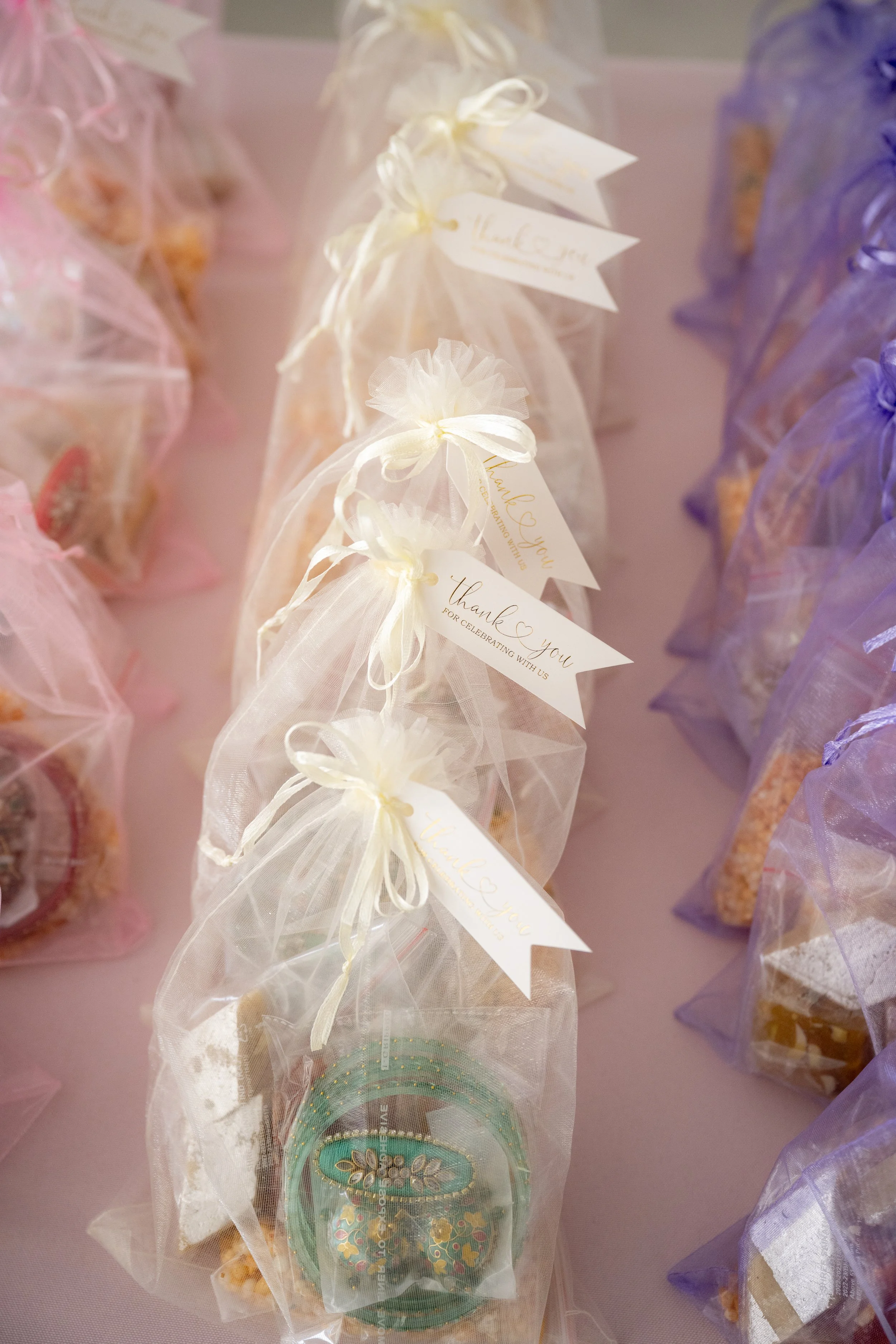 Clear plastic organza favor bags tied with ribbon, containing decorated cookies and tags that say 'Thank You for celebrating with us' and 'Kaleidoscope.' The background has pink and purple wrapped treats.
