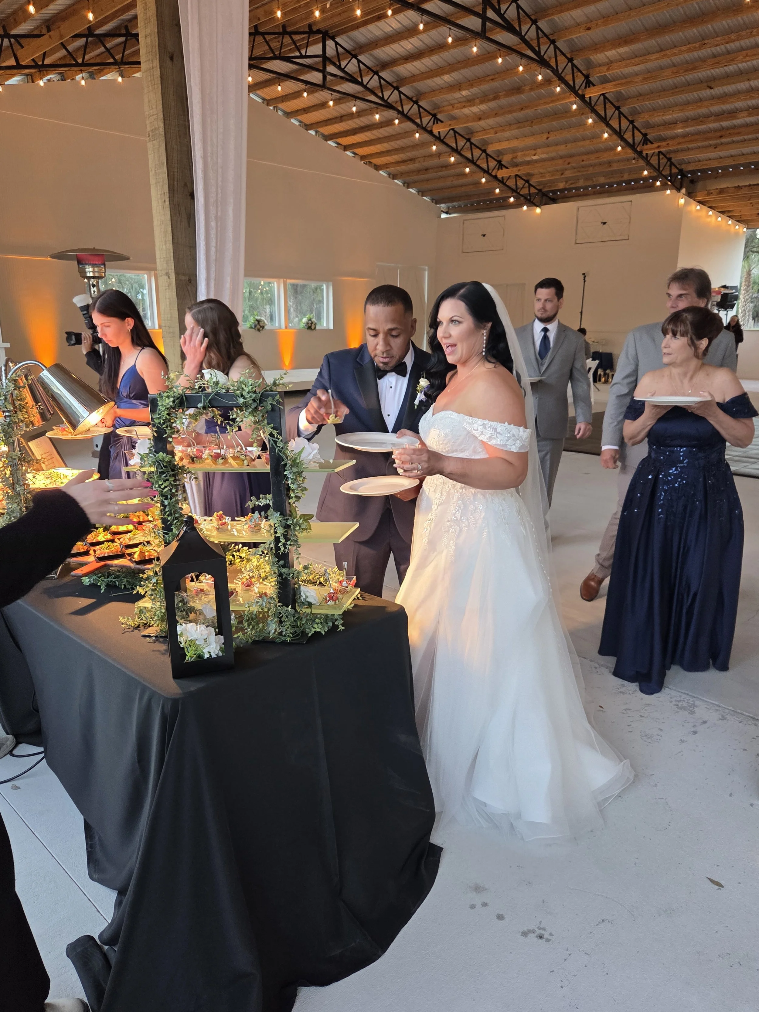 At a wedding reception, the bride and groom stand at a dessert table, serving themselves wedding cake. Guests are in the background, and the reception is decorated with string lights, with a rustic, open-beamed ceiling.