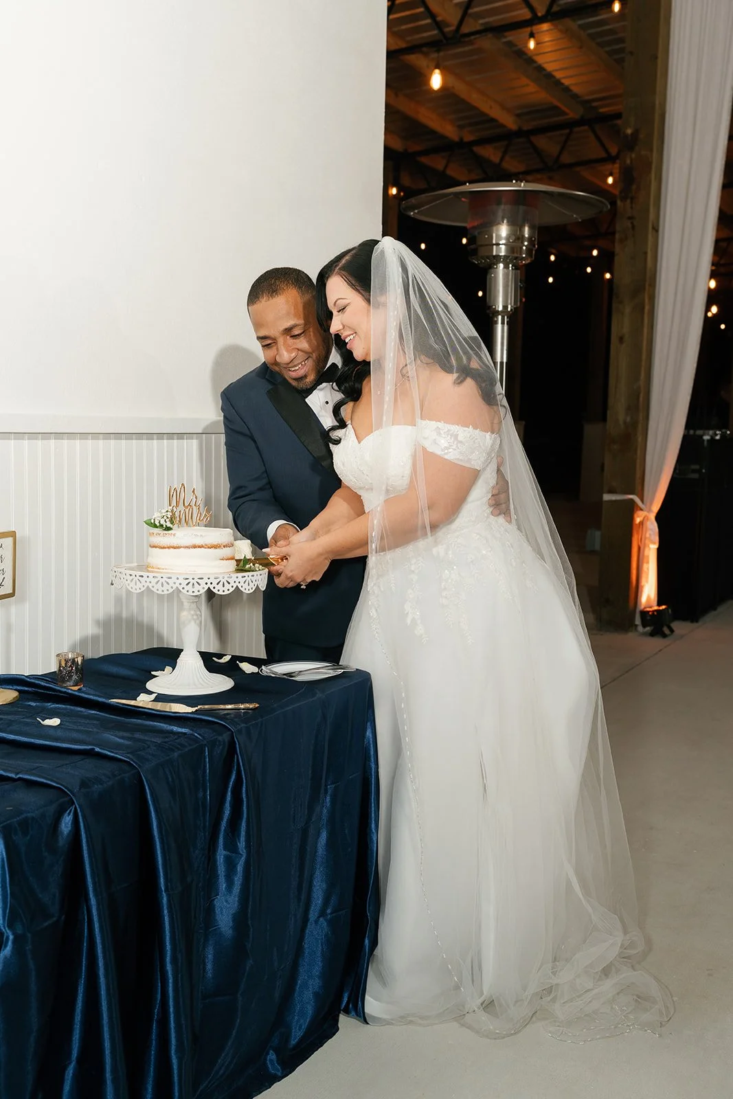 A bride and groom cutting a wedding cake together, smiling, with the bride wearing a white wedding dress and veil, and the groom in a navy tuxedo, at a wedding reception.