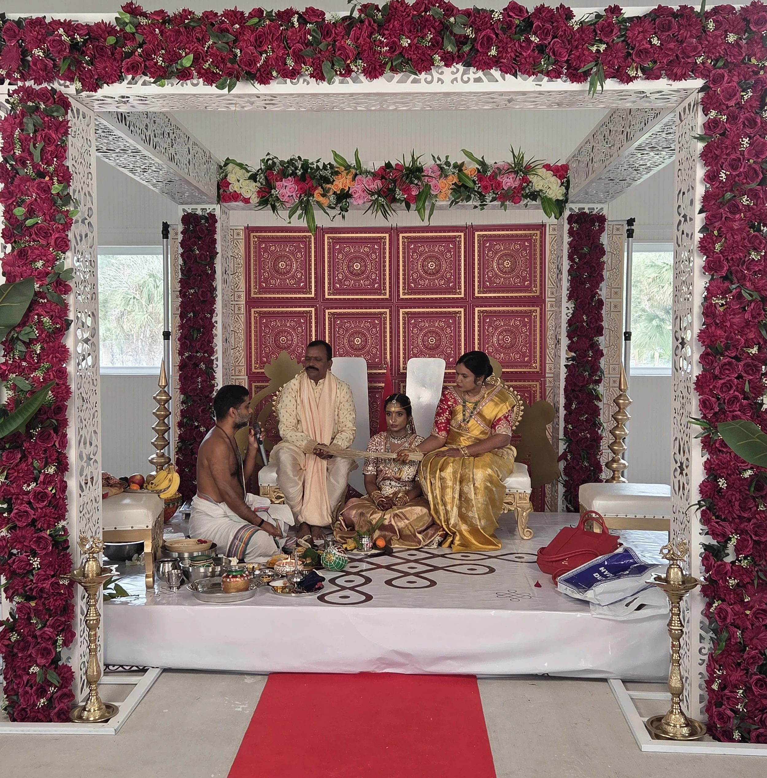 A wedding mandap decorated with red and white flowers, featuring intricate white lattice pillars, placed on a red carpet. Inside the mandap, people are seated during a traditional ceremony with offerings and wedding accessories visible.