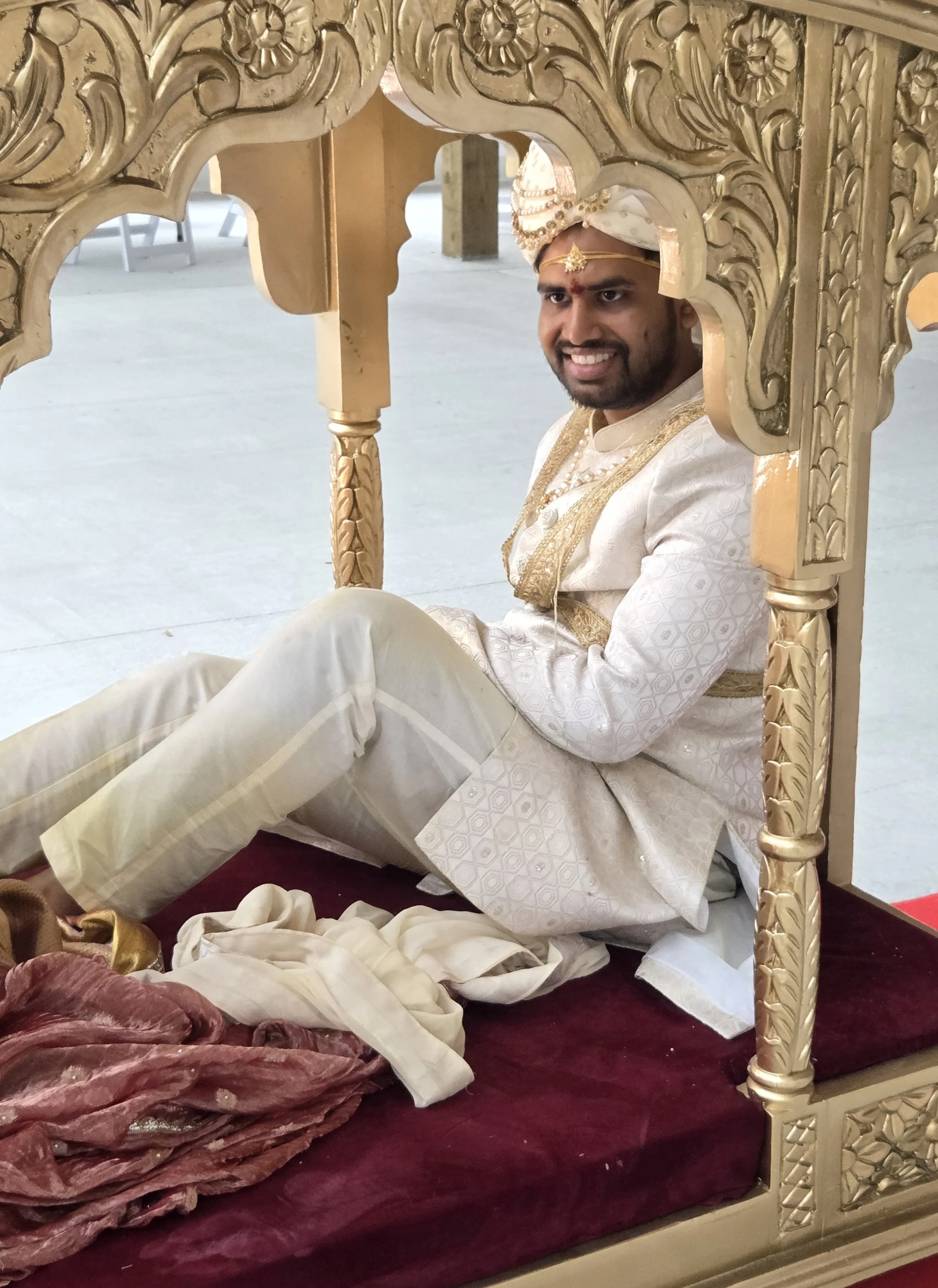 A man dressed in traditional Indian wedding attire, sitting on the floor under a decorated canopy, smiling. He wears a cream-colored embroidered sherwani, a turban with gold embellishments, and jewelry. The canopy has ornate gold carvings, and there 