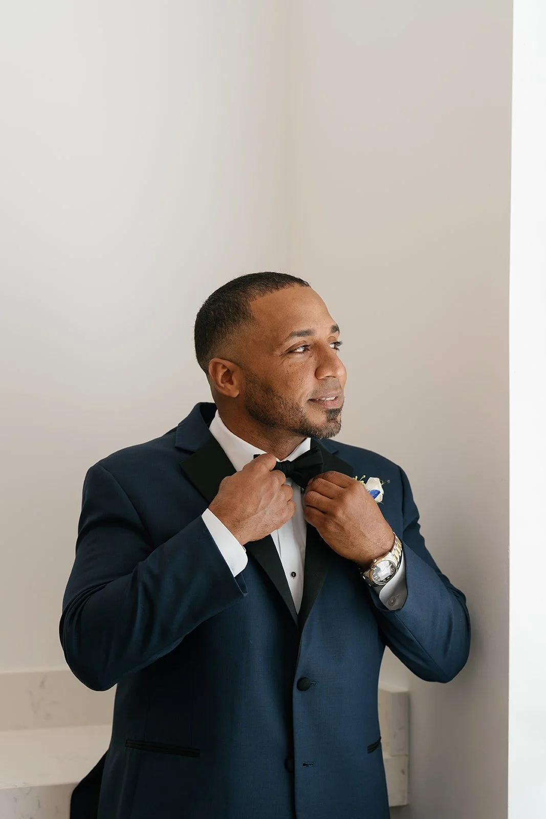 A man in a dark blue tuxedo adjusts his black bow tie in front of a plain white wall.