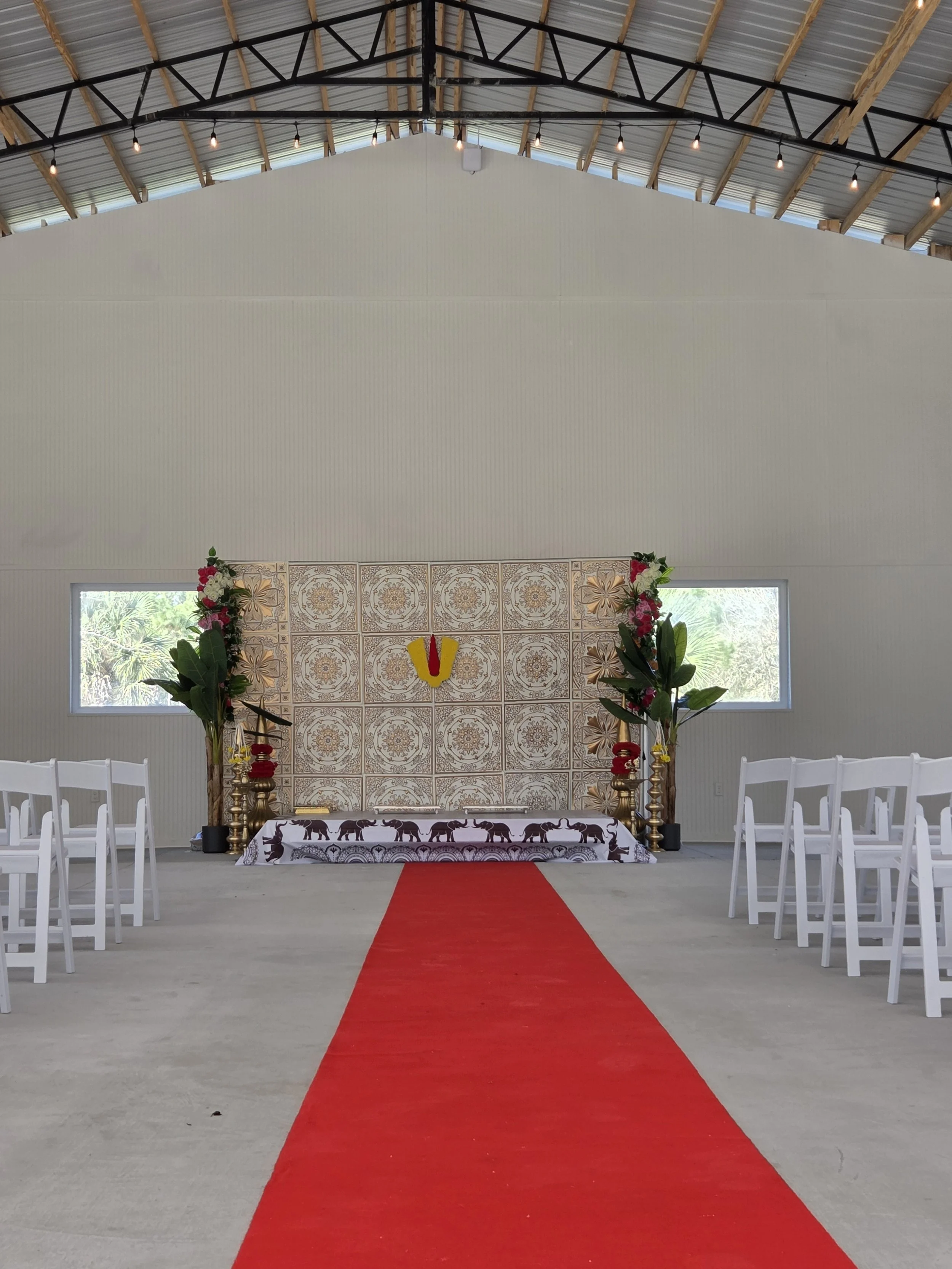 Indoor wedding ceremony setup with white chairs arranged on either side of a red carpet leading to an altar. The altar features a decorative back wall with patterns and floral arrangements with pink and white flowers, green leaves, and gold accents.