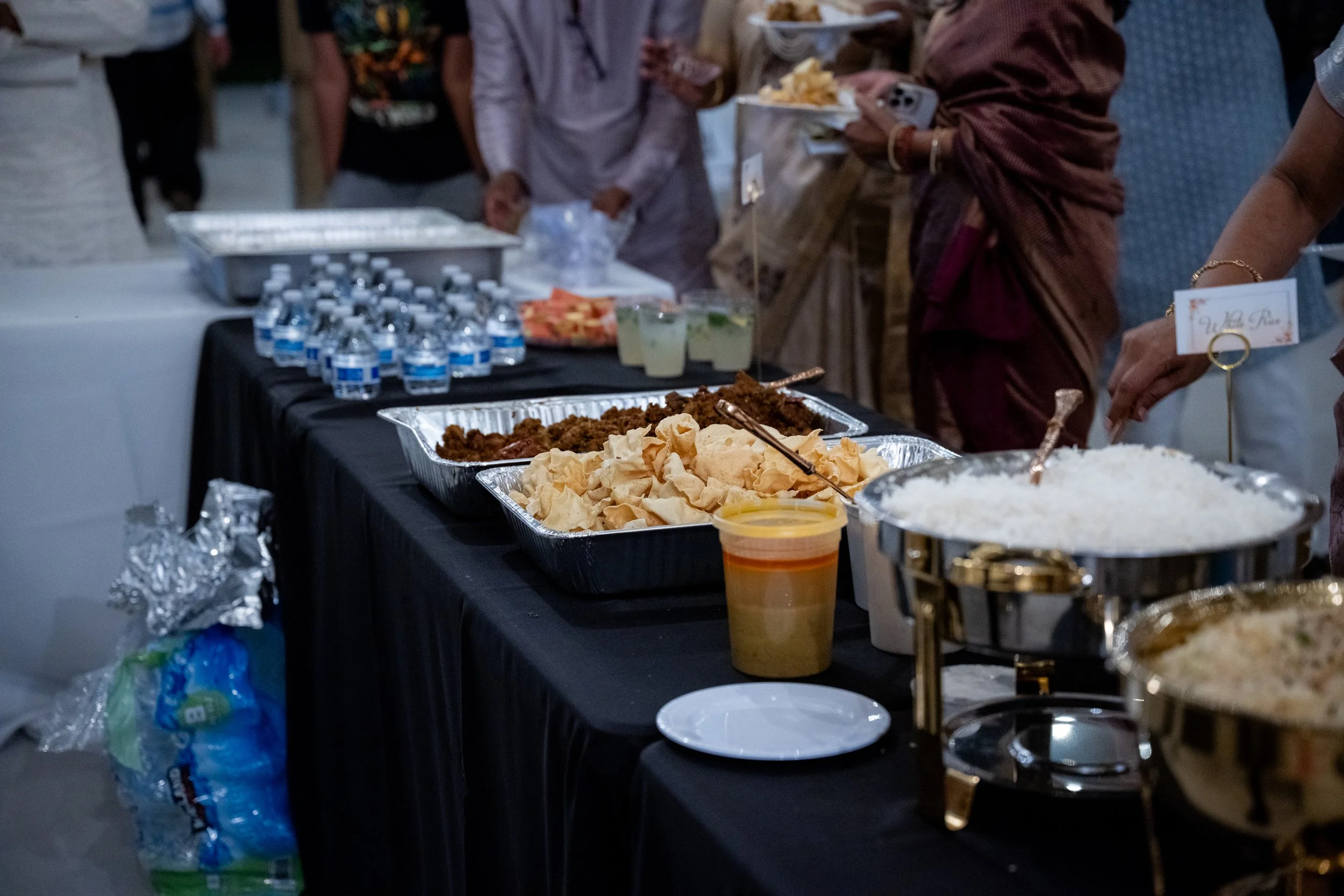A buffet table with various dishes including rice, fried food, chips, and drinks, with people serving themselves at a social gathering.