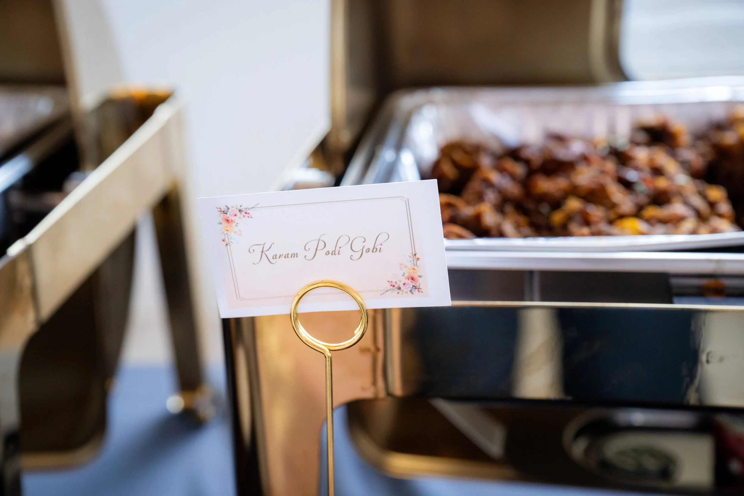 Close-up of a sign that says 'Karam Podi Gobi' with a floral design, placed on a metal stand near trays of food, possibly at a buffet or food display.
