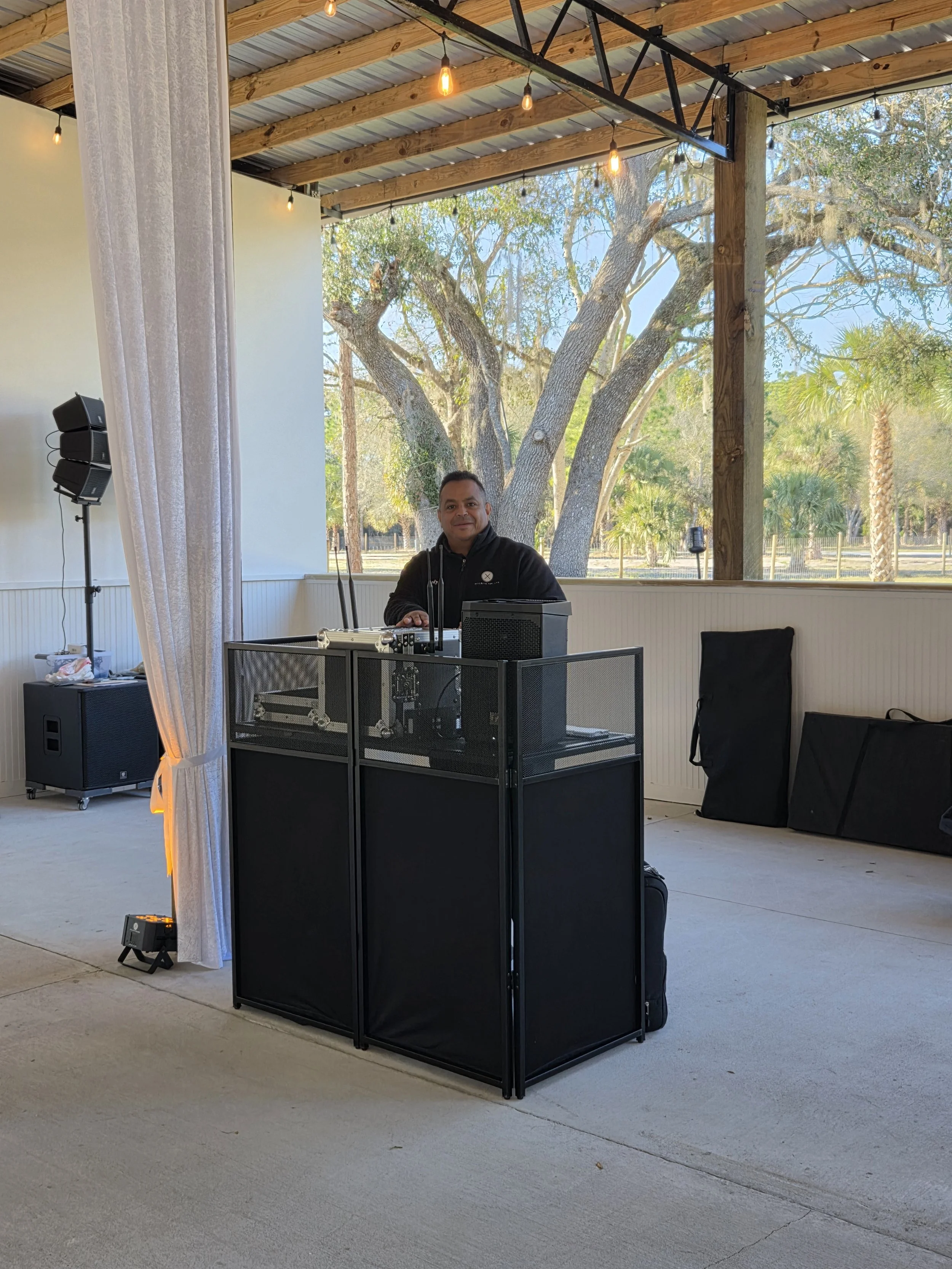 Man standing behind DJ table with outdoor background of trees and sky, enclosed by a metal and wood pavilion with string lights.