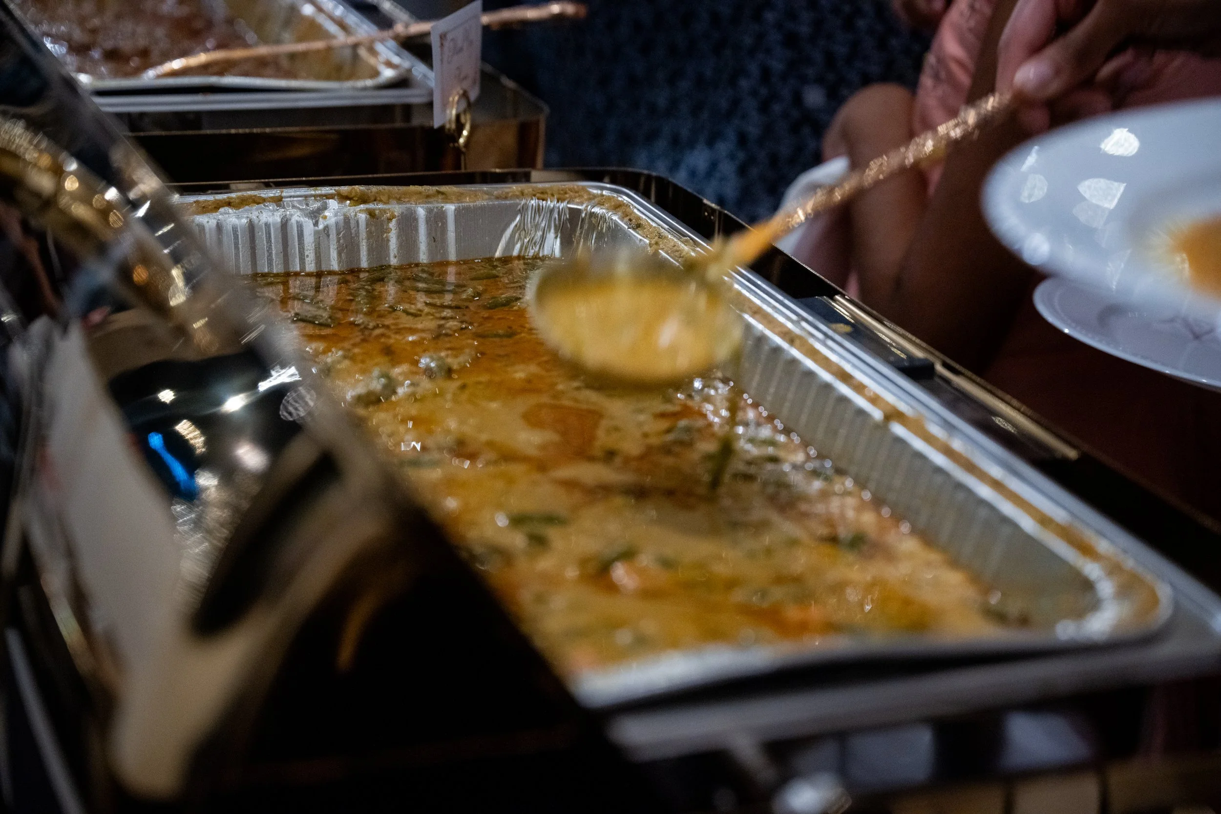 A kitchen or cafeteria setting with a person scooping from a large aluminum tray of food, possibly casserole or stew, with a white plate in hand.