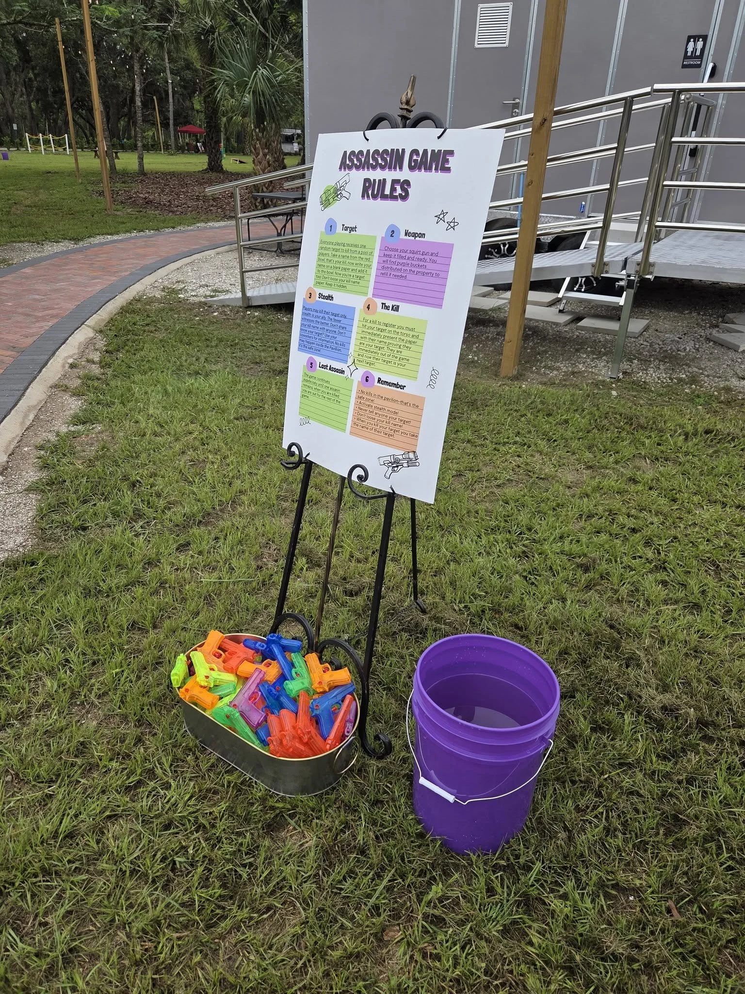 Sign displaying rules for a game called Assassin, with buckets of toy water guns and empty buckets nearby, set outdoors on a grassy area near a building with a ramp.