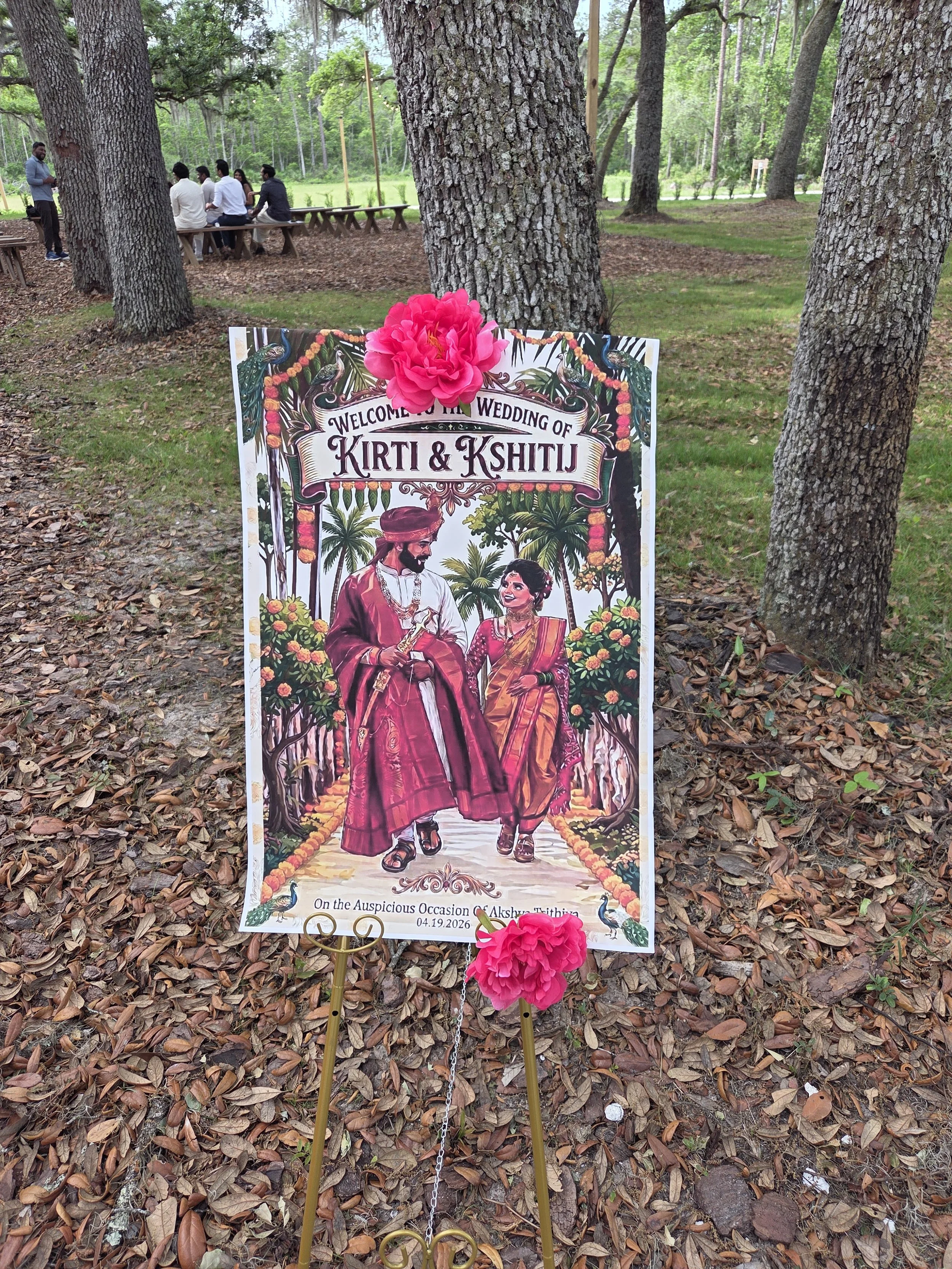 A wedding sign decorated with pink flowers featuring illustrated Indian groom and bride, with text welcoming guests, set outdoors among trees and fallen leaves.