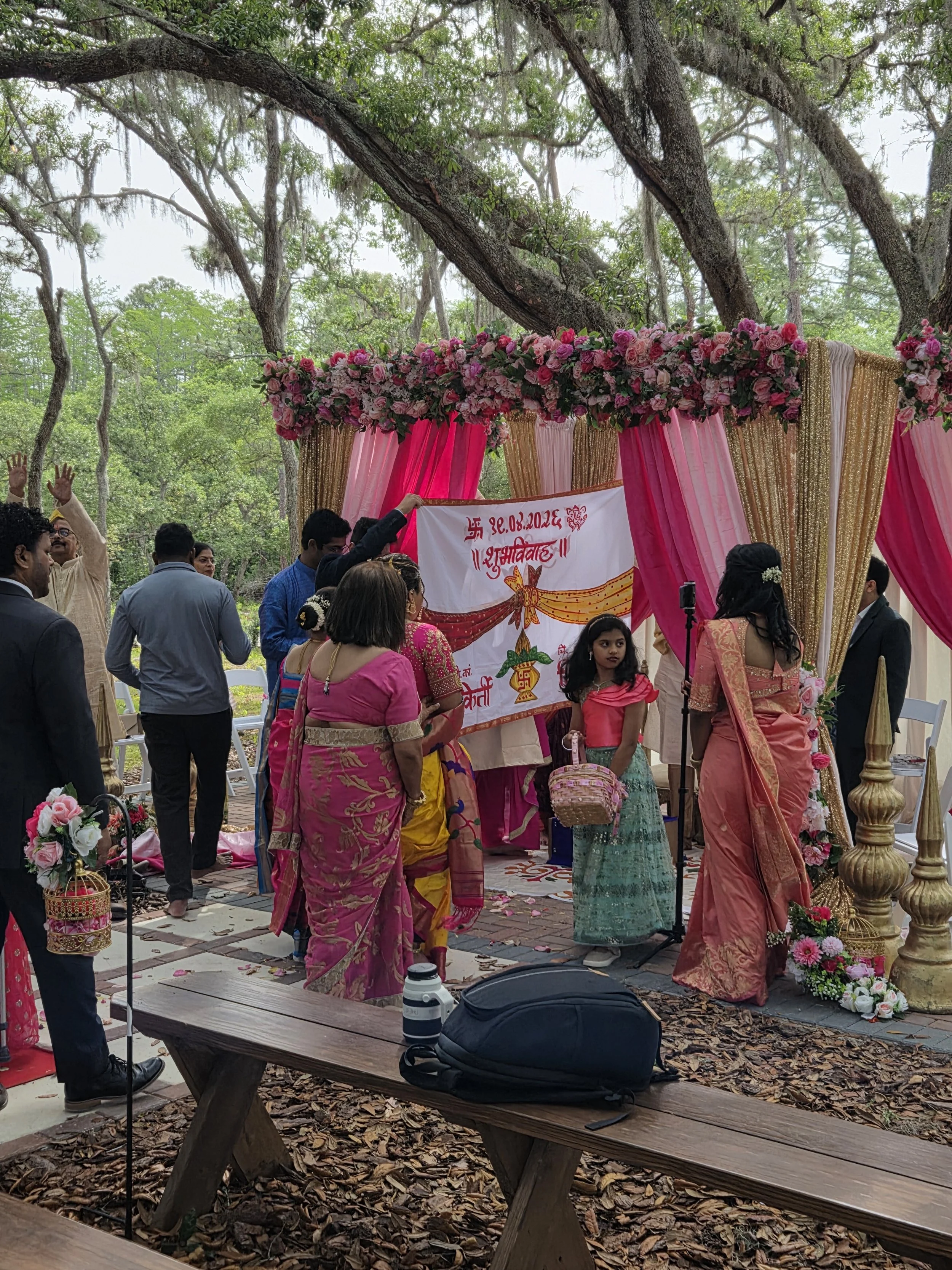 A group of people gathered under a decorated canopy with pink and gold drapes and floral arrangements, participating in a traditional Hindu wedding ceremony outdoors surrounded by trees.