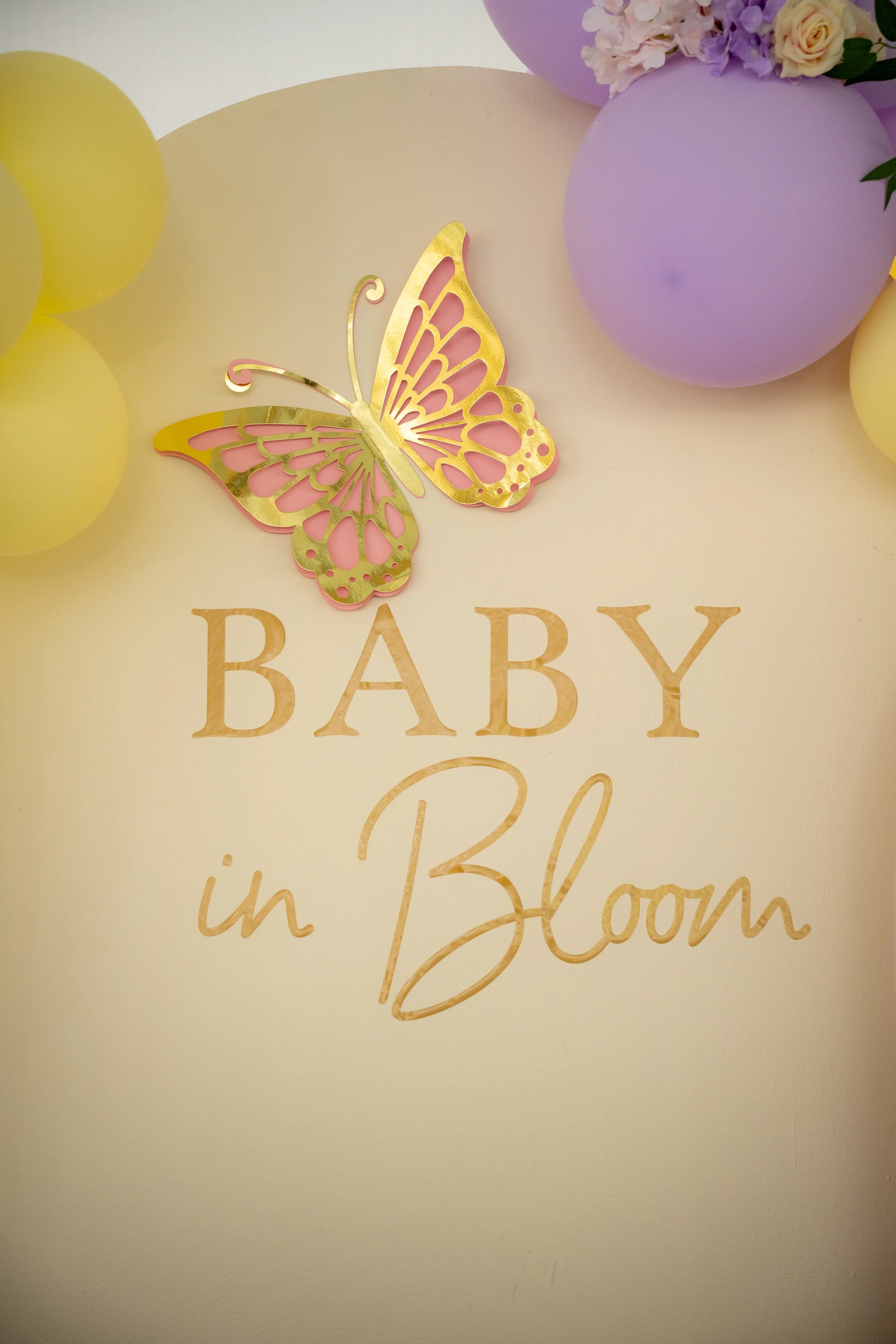 Decorative sign with gold butterfly and pastel-colored balloons in purple and yellow, with floral arrangements, celebrating the arrival of a baby in Bloom.