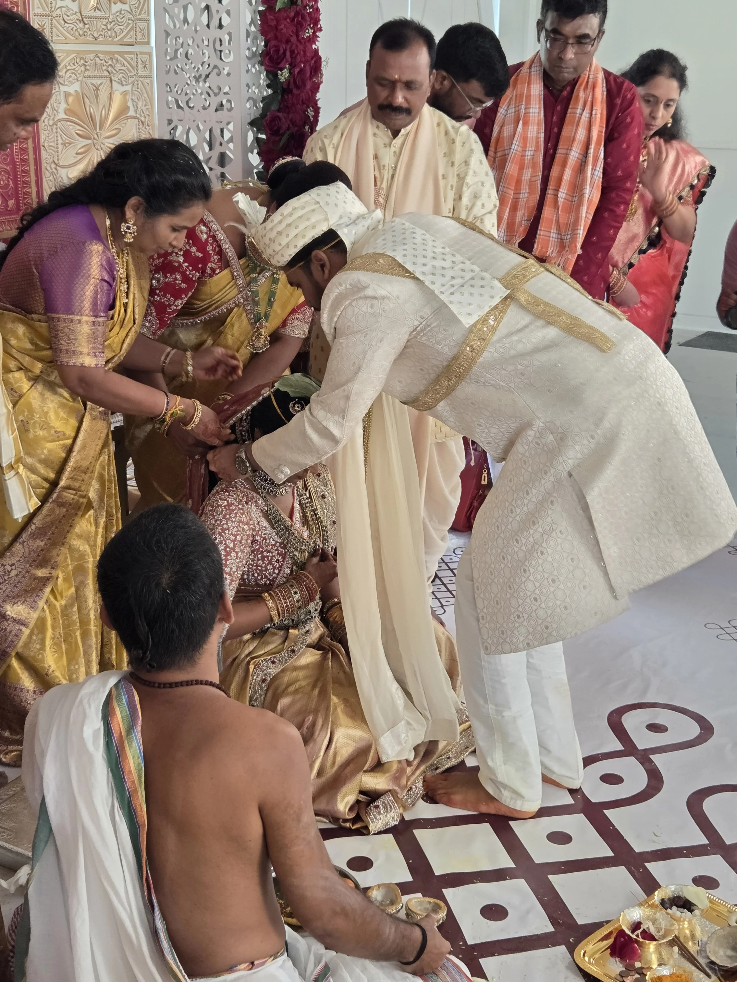 A traditional Hindu wedding ceremony taking place indoors with several people dressed in colorful, ornate attire bowing and performing rituals. The groom, dressed in white, is bending forward while the bride, sitting in front, is adorned in jewelry a