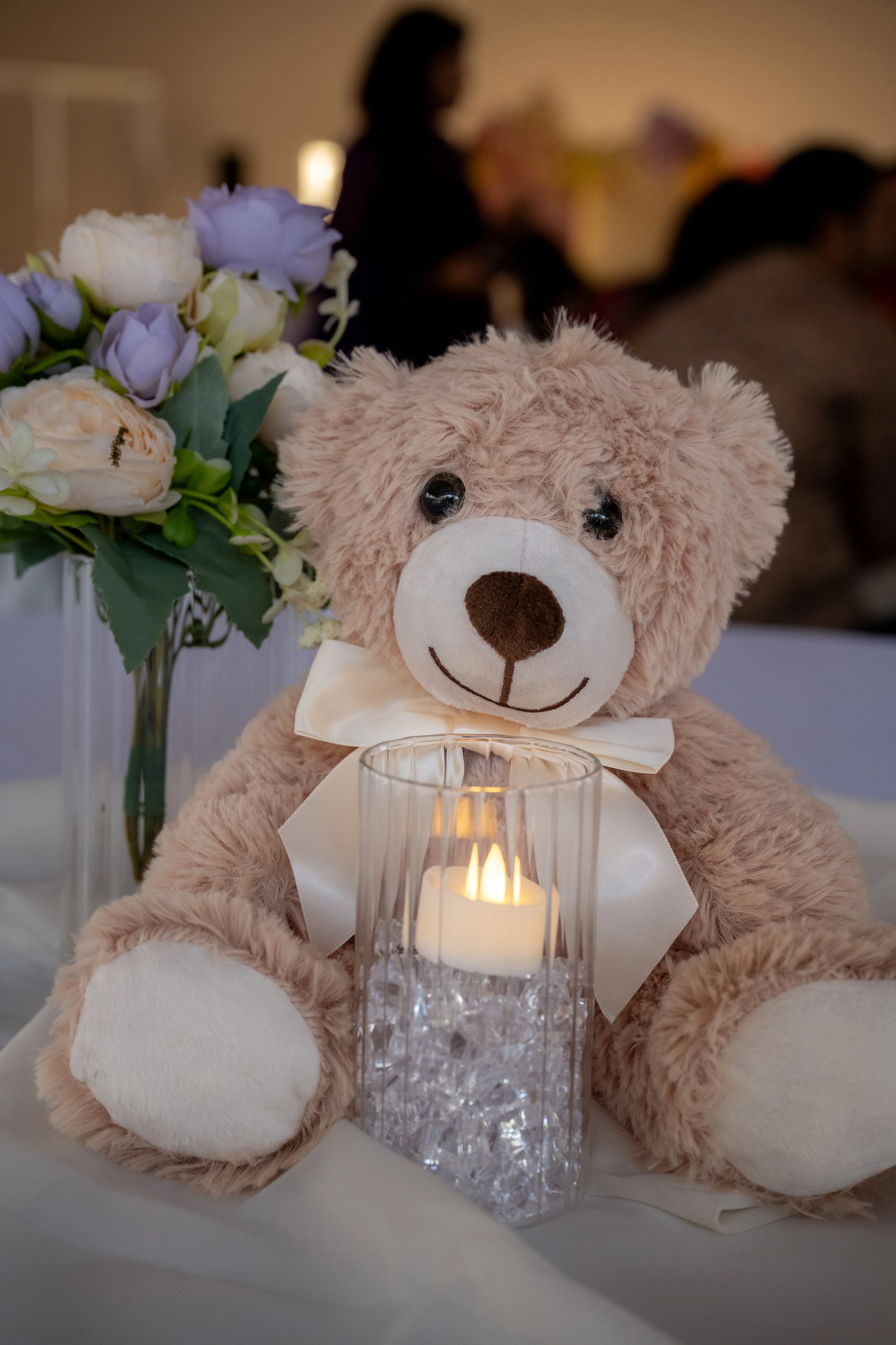 A close-up of a teddy bear with a cream-colored bow, sitting in front of a lit candle in a glass holder. In the background, there is a blurred bouquet of pale purple and cream roses and an arrangement of flowers.