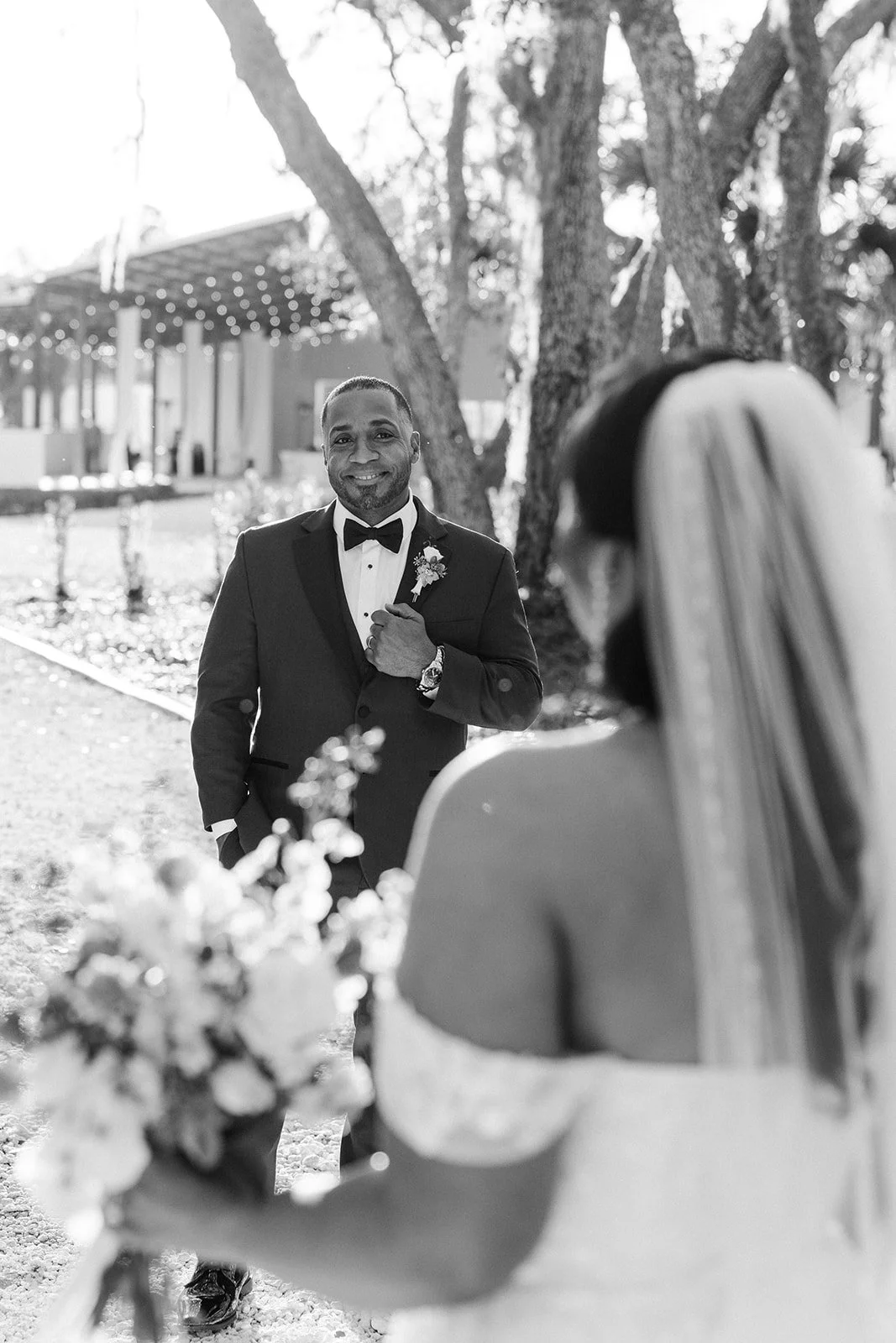 A groom in a tuxedo with a boutonniere and bow tie smiling while looking at his bride, who is holding a bouquet of flowers, outdoors in a park-like setting with trees and string lights in the background.