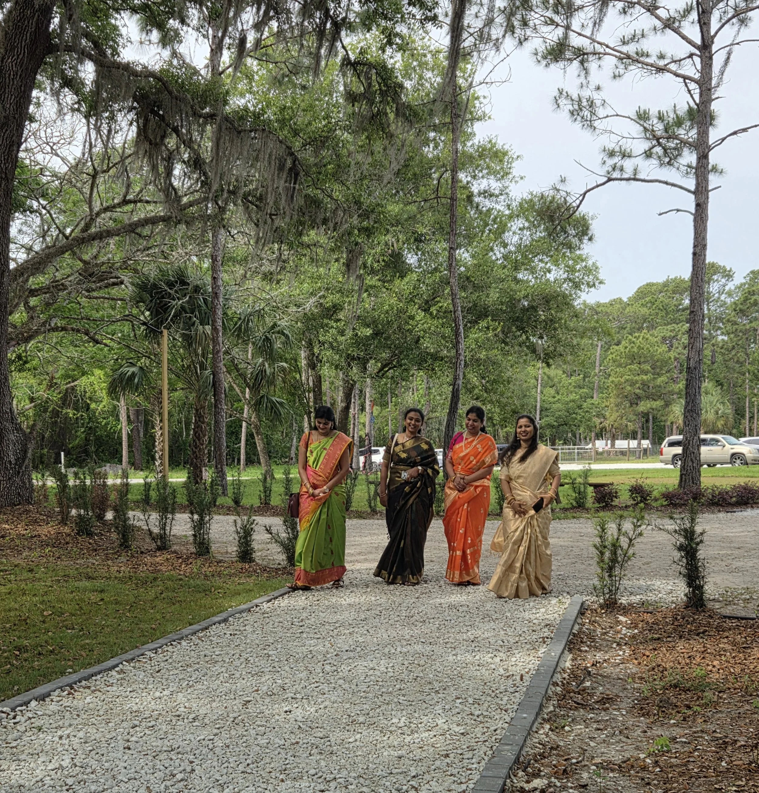 Four women dressed in traditional Indian sarees walking on a gravel path in a park with tall trees and greenery.