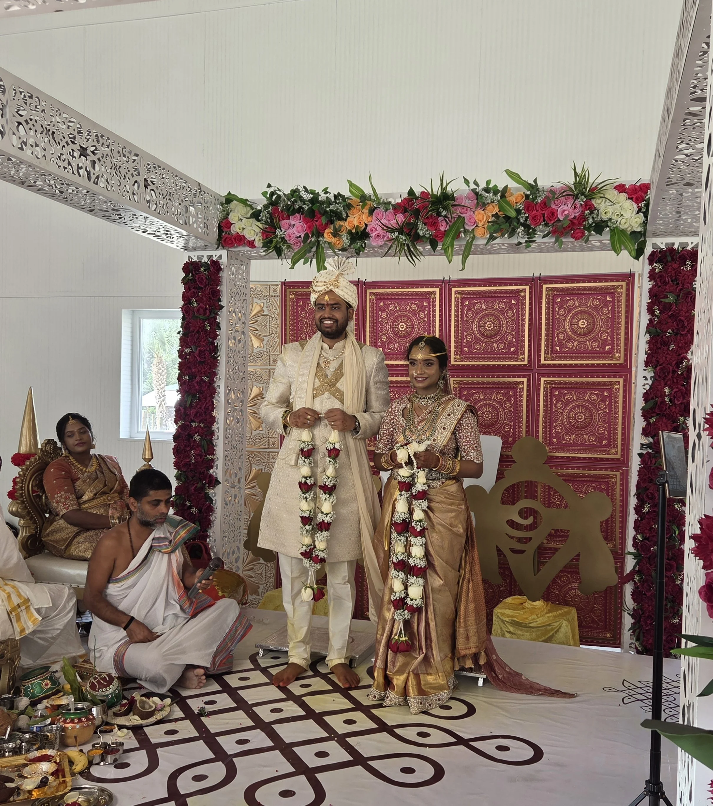 A traditional Indian wedding ceremony with a bride and groom standing under a decorated floral canopy, with priests and attendees seated nearby.