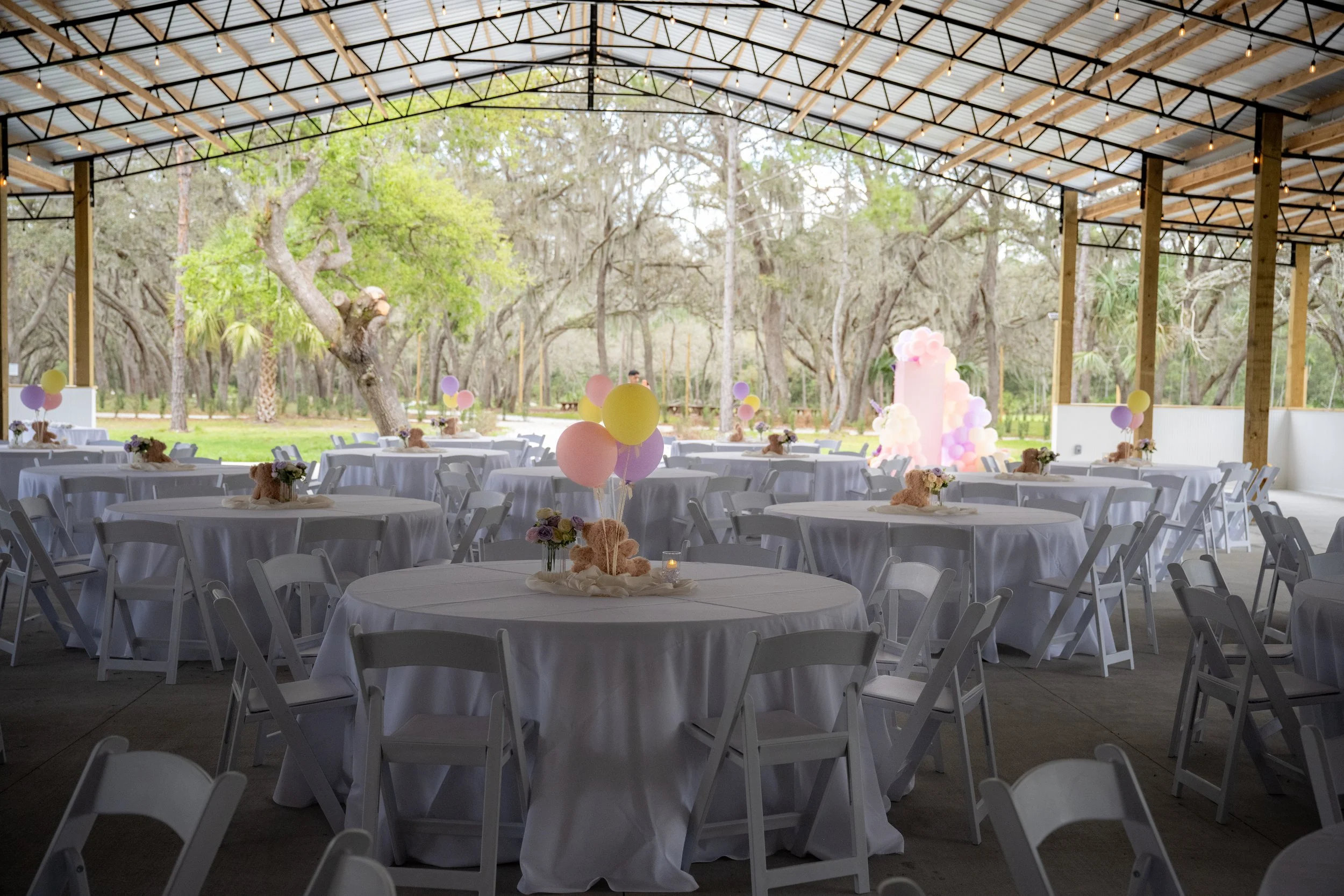 Indoor venue with round tables covered in white tablecloths, decorated with teddy bear centerpieces, surrounded by white folding chairs, balloons in pastel colors, and an outdoor view of trees with a canopy overhead, Bonfire styled balloon display, a