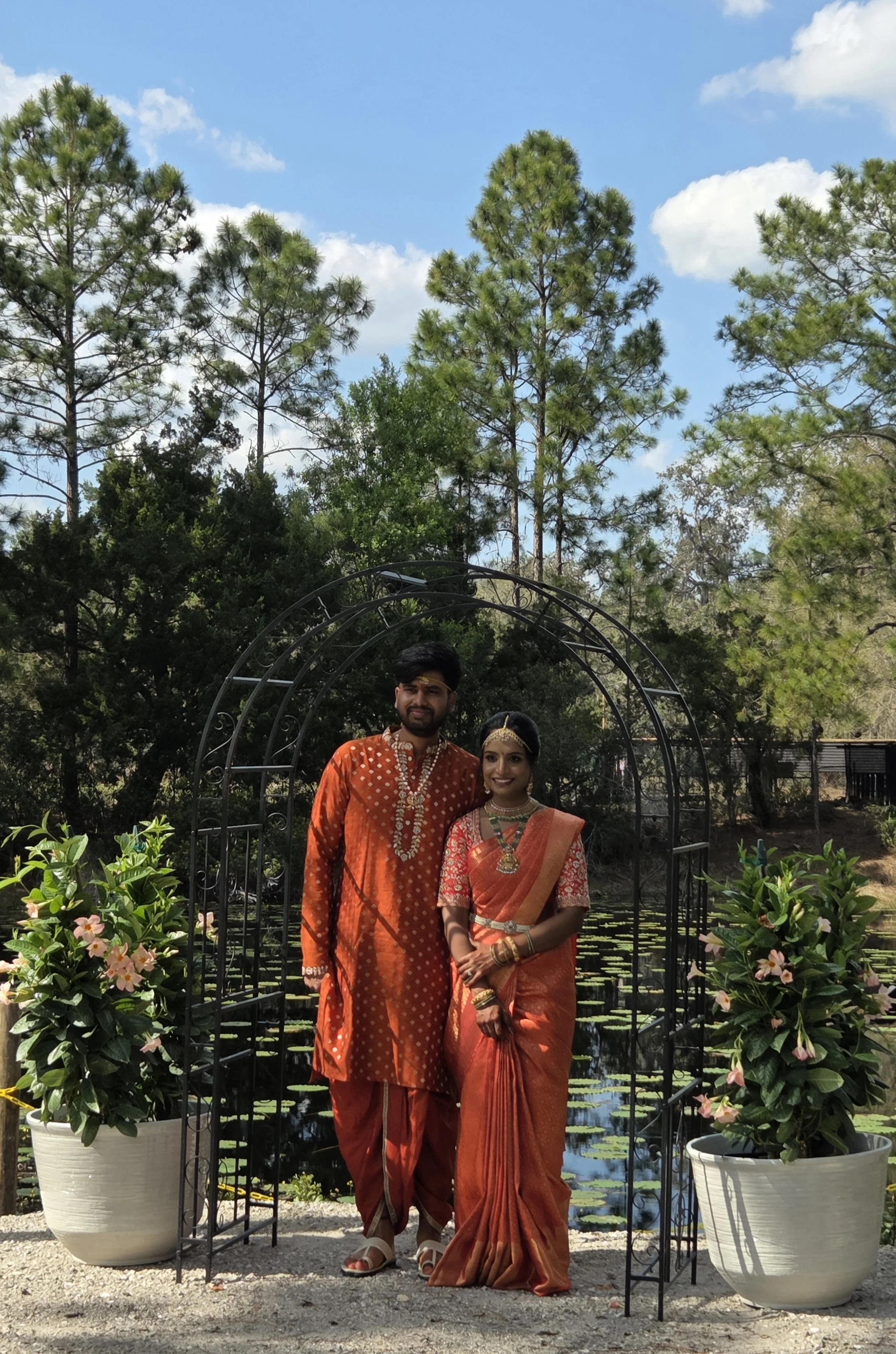 A man and woman dressed in traditional Indian attire standing under a metal archway with greenery and potted plants around, near a pond with water lilies, trees, and a blue sky with clouds in the background.