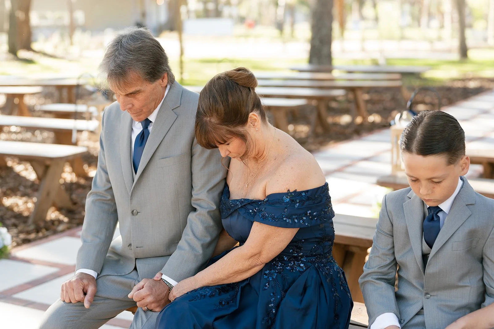Three people sitting on benches with heads bowed and eyes closed outdoors, two males in grey suits and a woman in a blue dress, in a serene setting with trees and picnic tables.