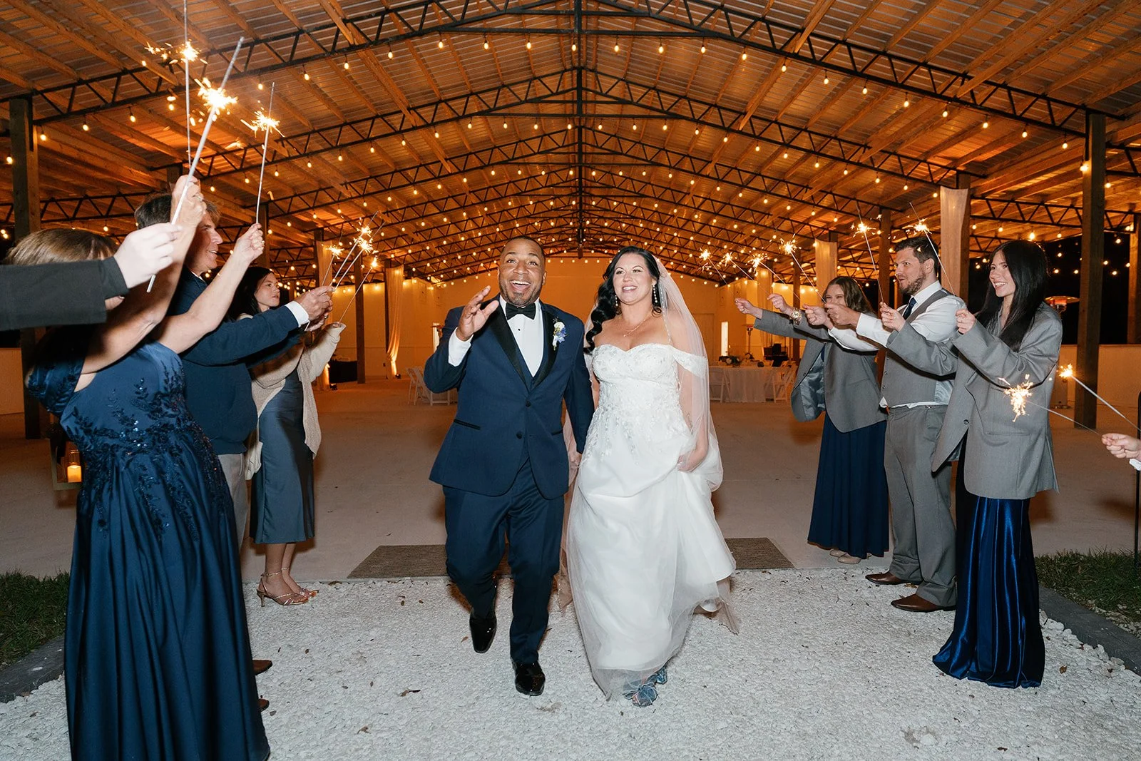 A newlywed couple walking through friends and family holding sparklers during a wedding reception indoors with wooden beams and string lights.