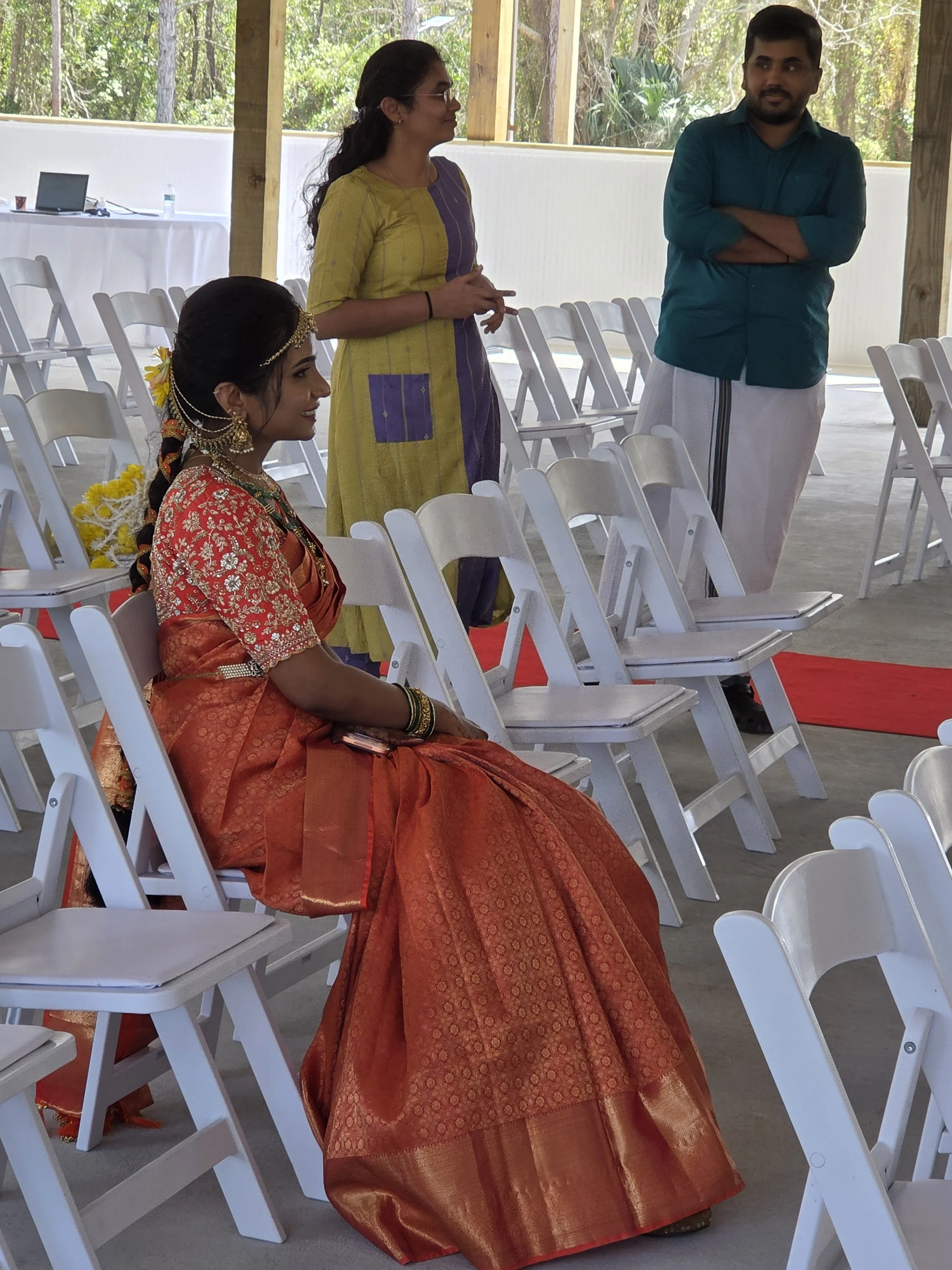 A woman dressed in traditional Indian attire with jewelry and flowers in her hair, sitting on a white chair, and two people standing nearby, one woman in a yellow and purple dress and one man in a teal shirt and traditional white pants, in an outdoor