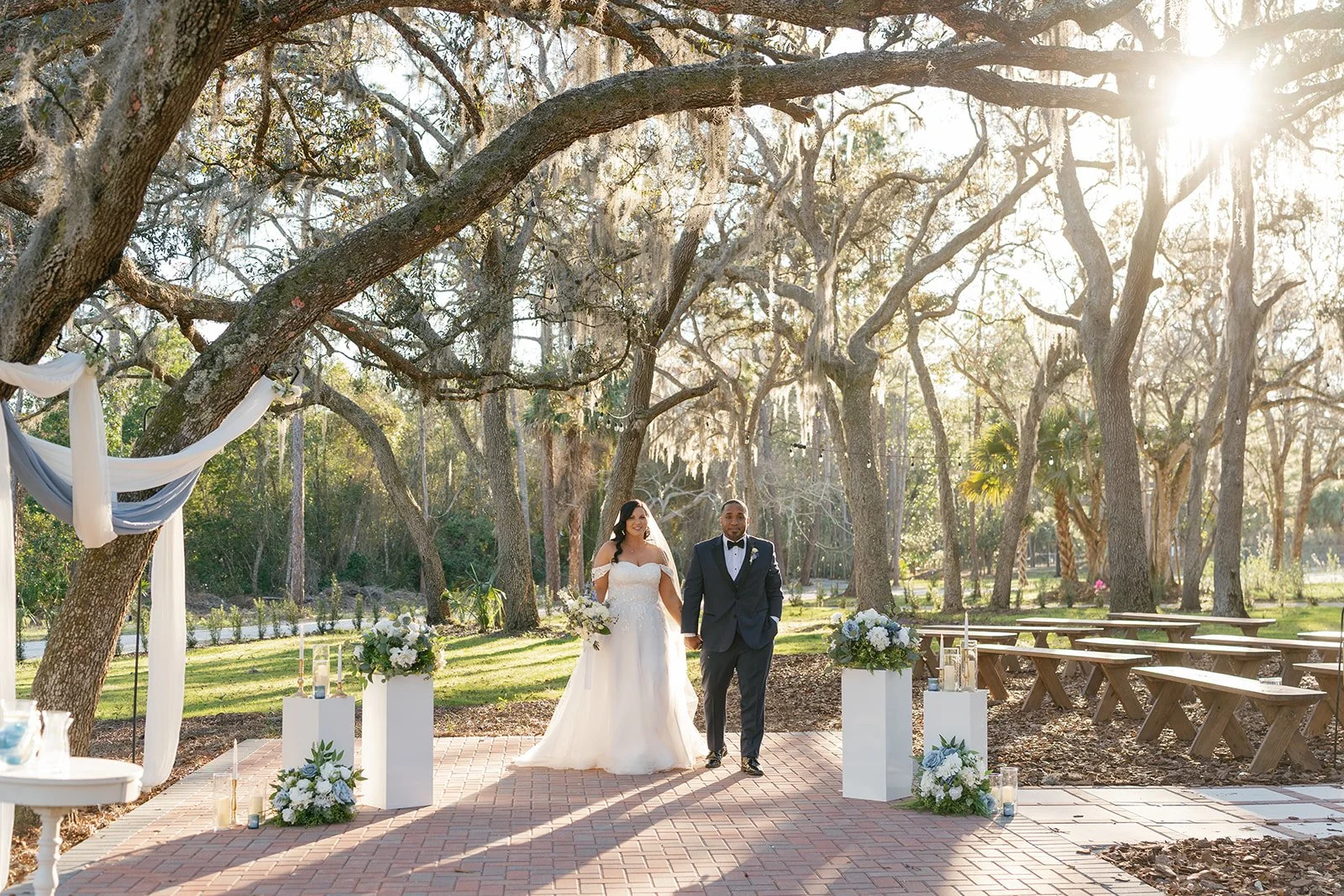 A bride and groom walking hand in hand outdoors during a wedding ceremony in a wooded area with sunlight filtering through the trees. The bride is wearing a white wedding gown and holding a bouquet, while the groom is dressed in a dark suit with a bo