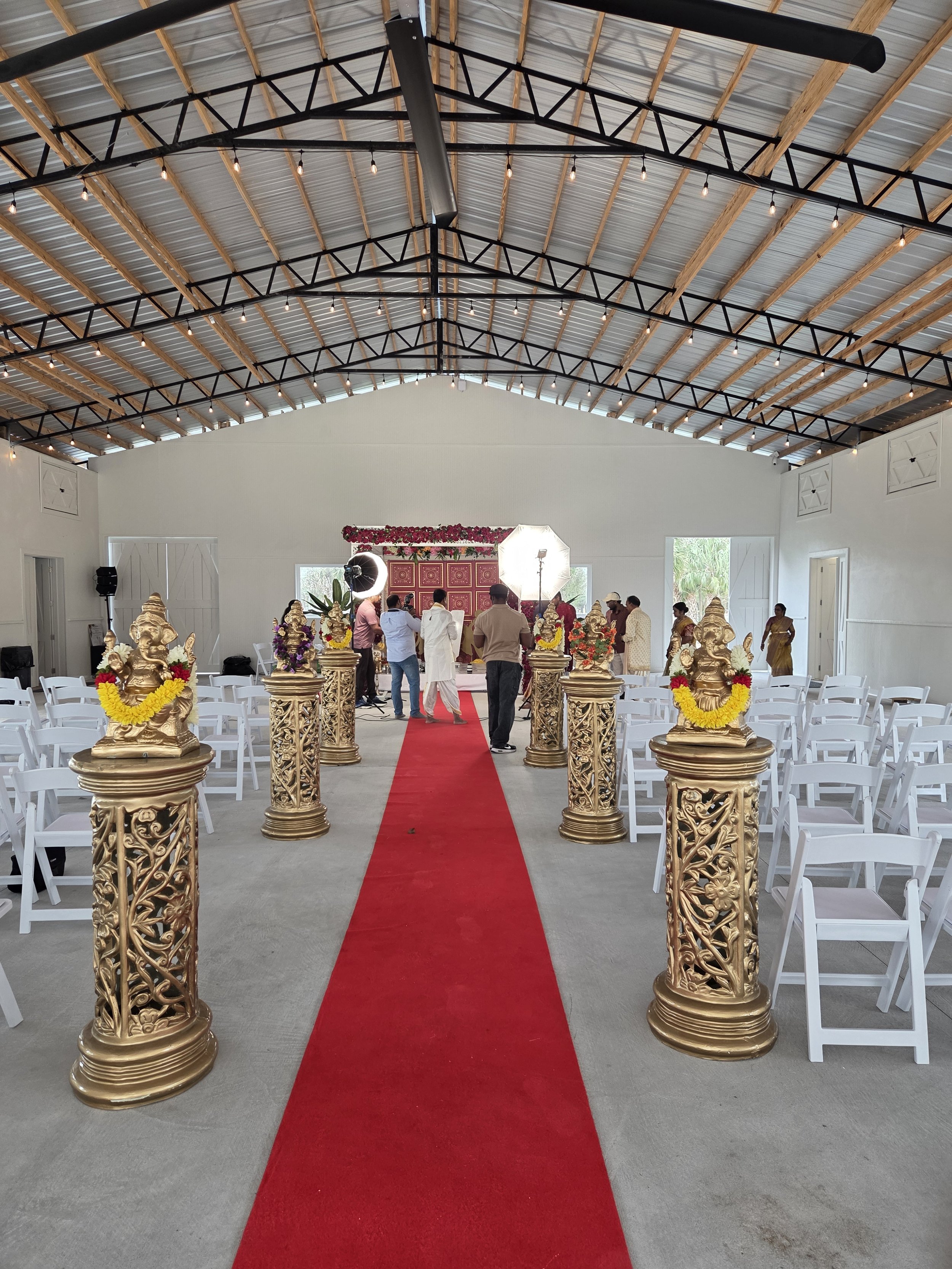 Interior of a decorated hall with a red carpet aisle, white chairs on either side, ornate gold statues, a backdrop with flowers, and a few people preparing for a ceremony, under a high ceiling with metal and wooden beams and string lights.