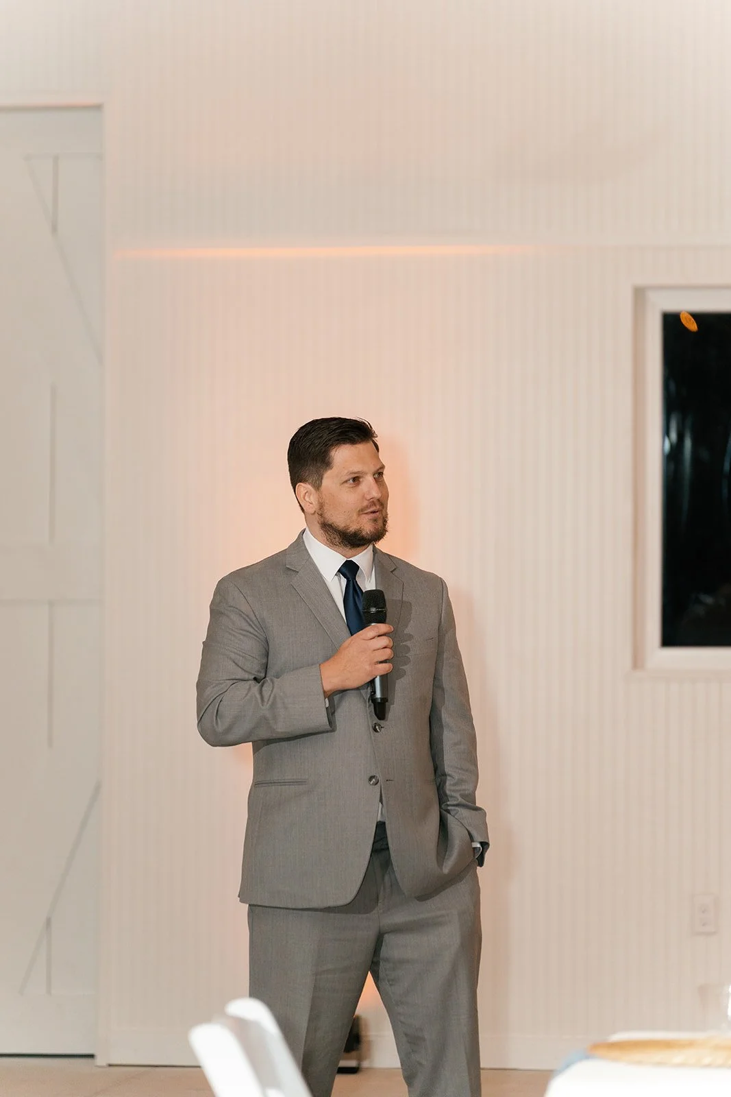 A man in a gray suit holding a microphone stands at a presentation or event, with a white wall and a window behind him.